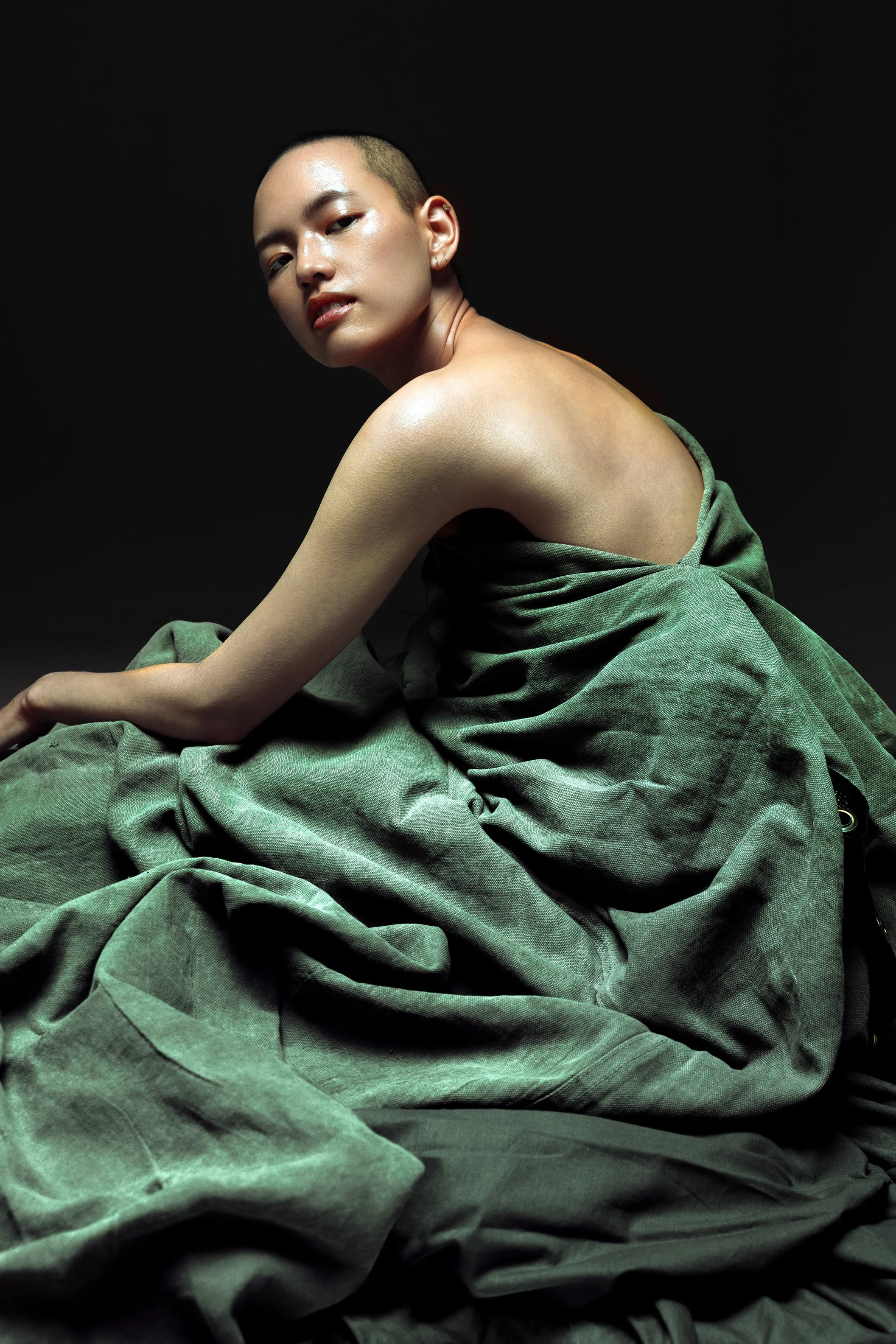 A person with a shaved head, sitting on green fabric, with no top, against a black background.