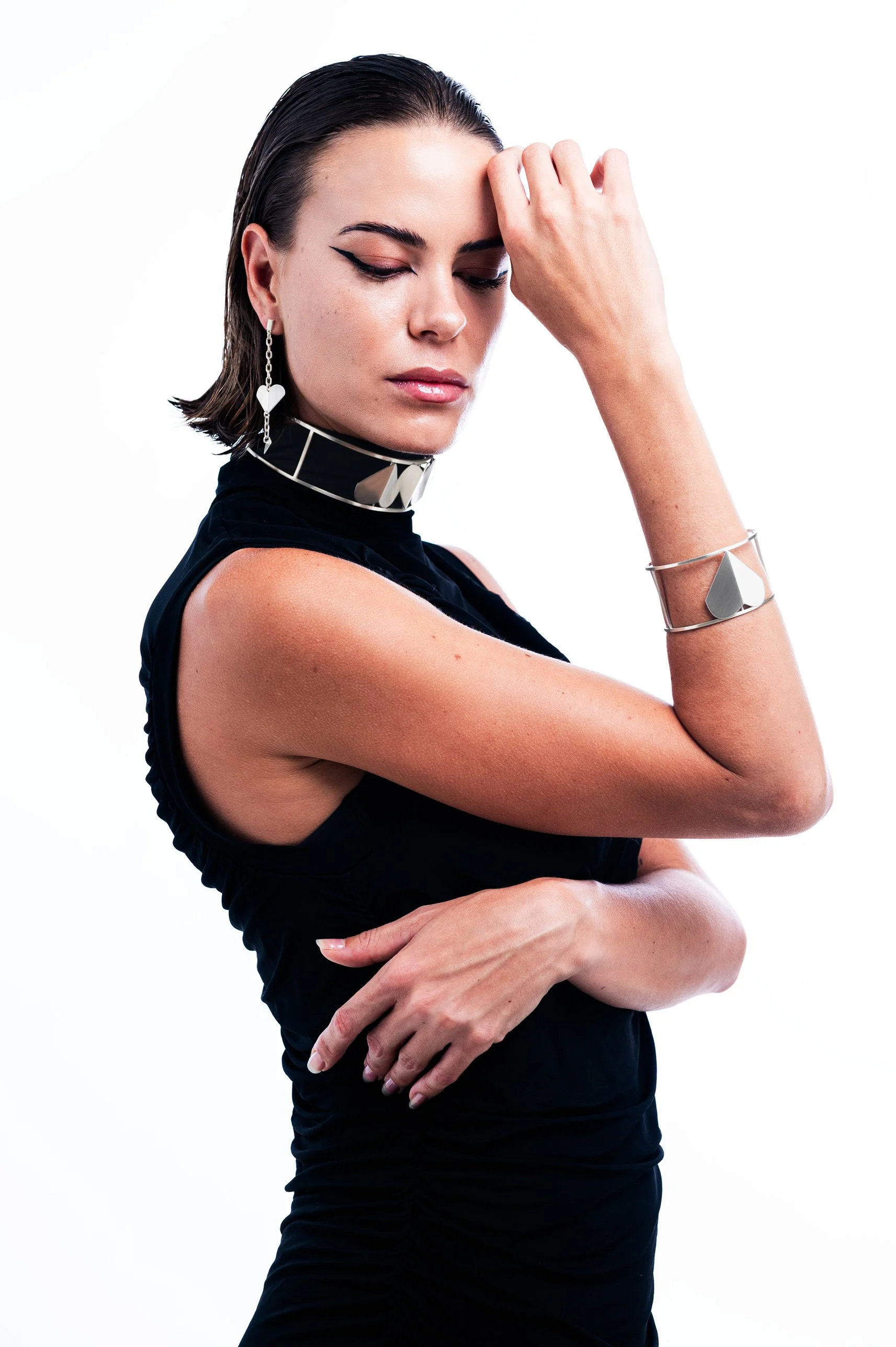 A woman with dark hair, styled slicked back, wearing a black sleeveless top and silver jewelry, including earrings, a choker, and bracelets, poses with her eyes closed against a white background.