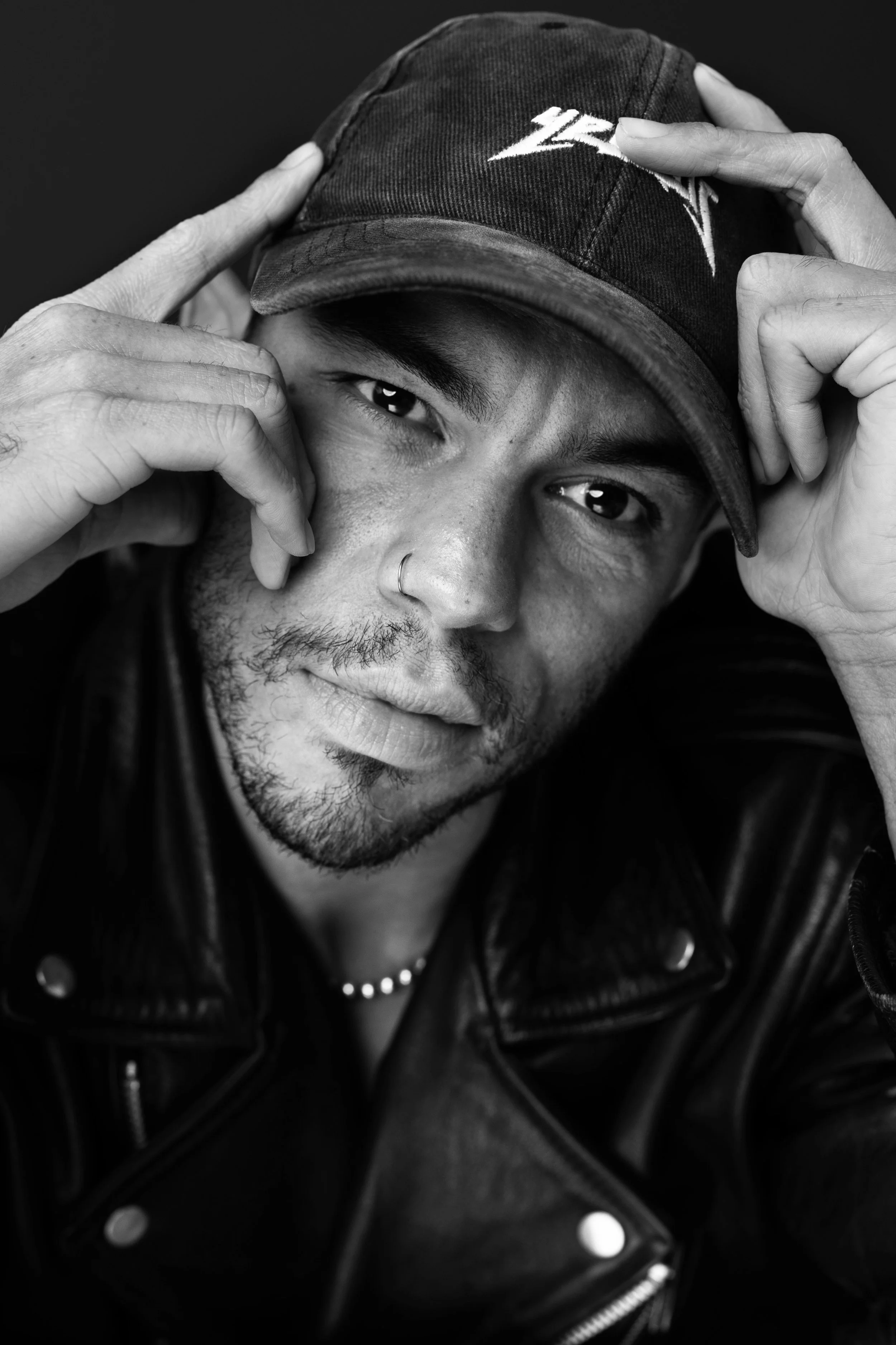 A young man with a nose ring, beard, and mustache wearing a leather jacket and a cap, looking at the camera in black and white.