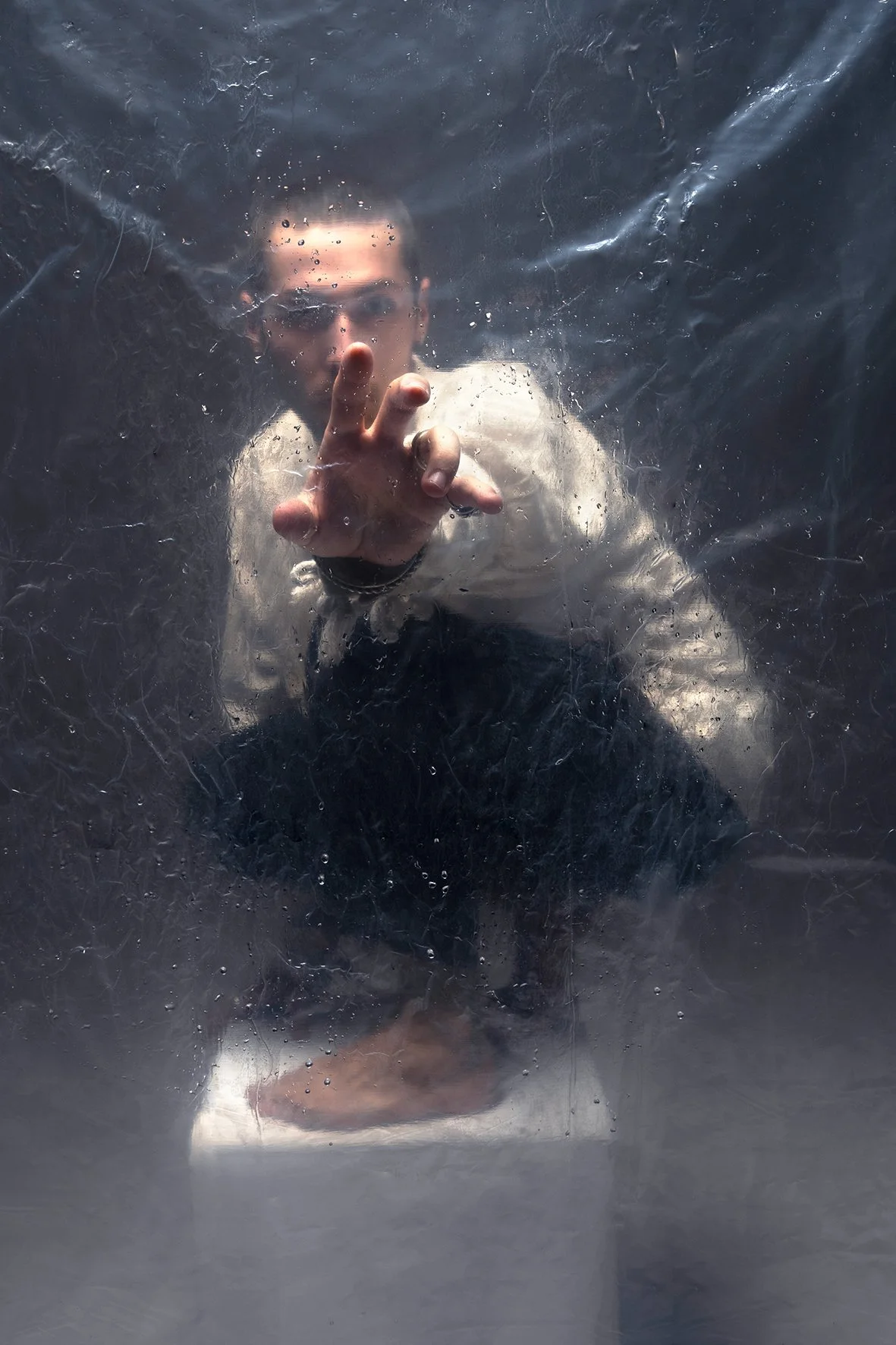 Person pointing forward through a foggy glass or plastic surface with water droplets, wearing glasses and a beige jacket.
