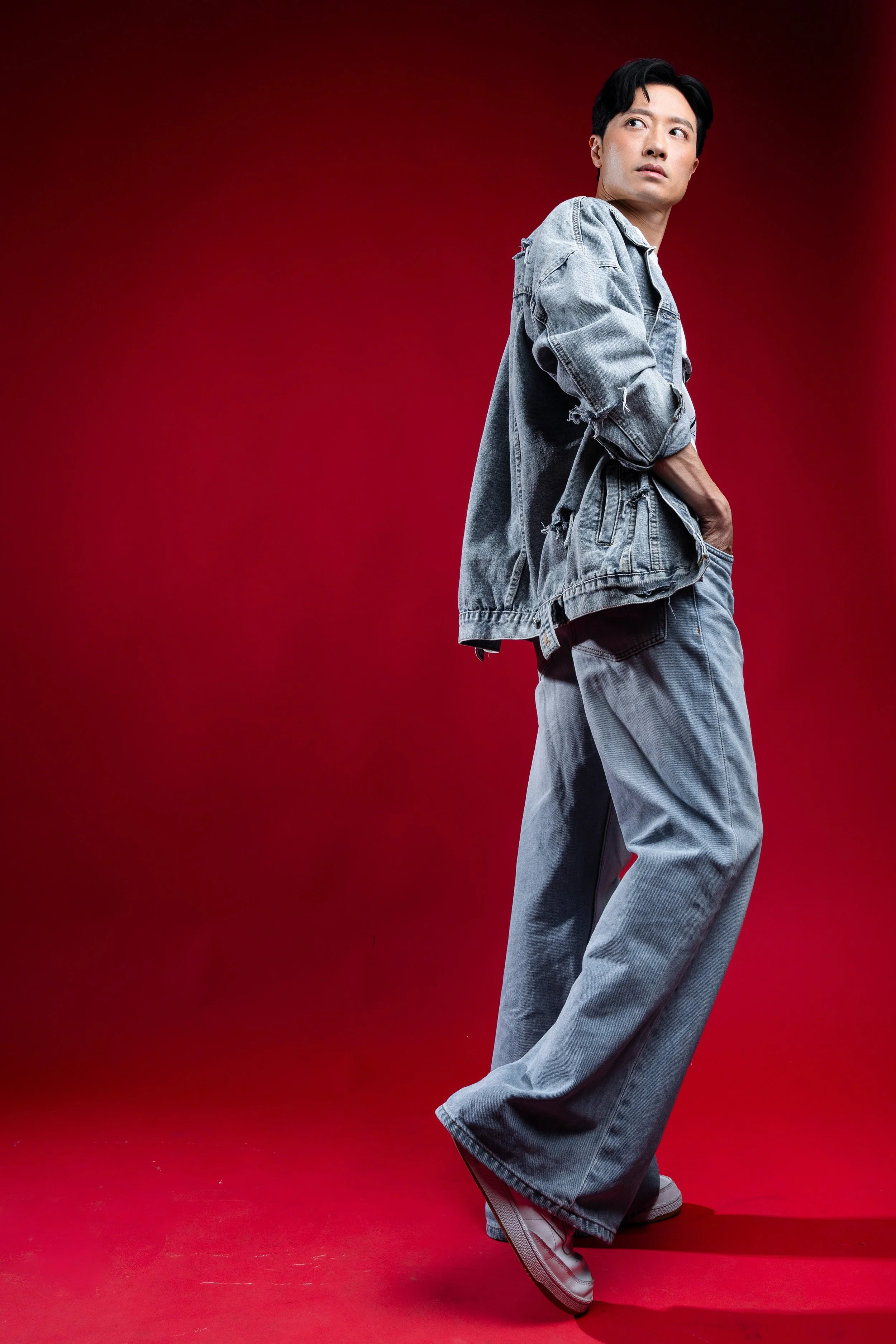 A young man in a distressed denim jacket and loose denim jeans strikes a pose against a red background.
