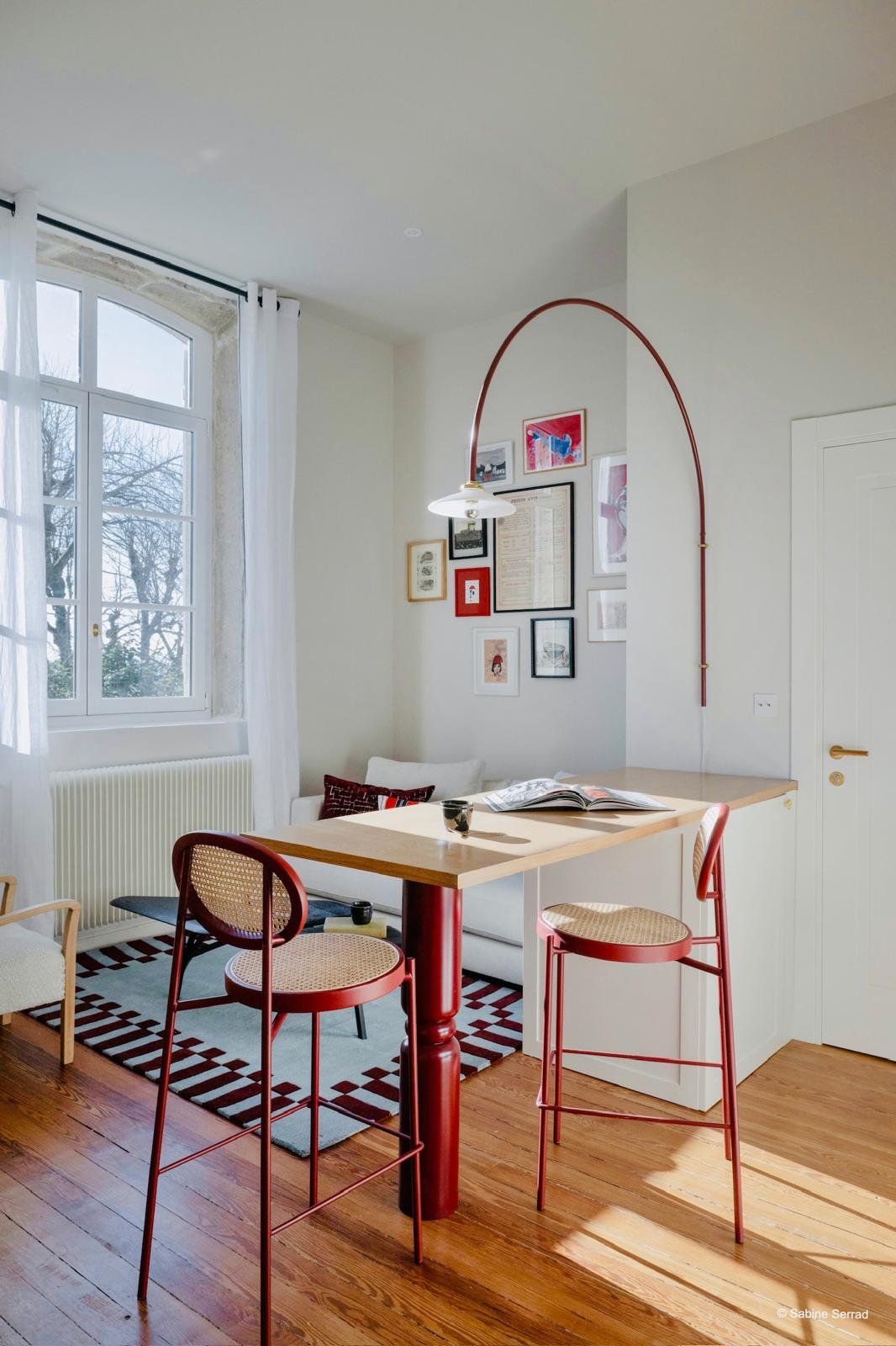 Intérieur d'une cuisine ou salle à manger lumineuse avec une table en bois, deux chaises rouges, un mur avec des cadres d'art, une fenêtre avec rideaux blancs, et un plan de travail en bois.