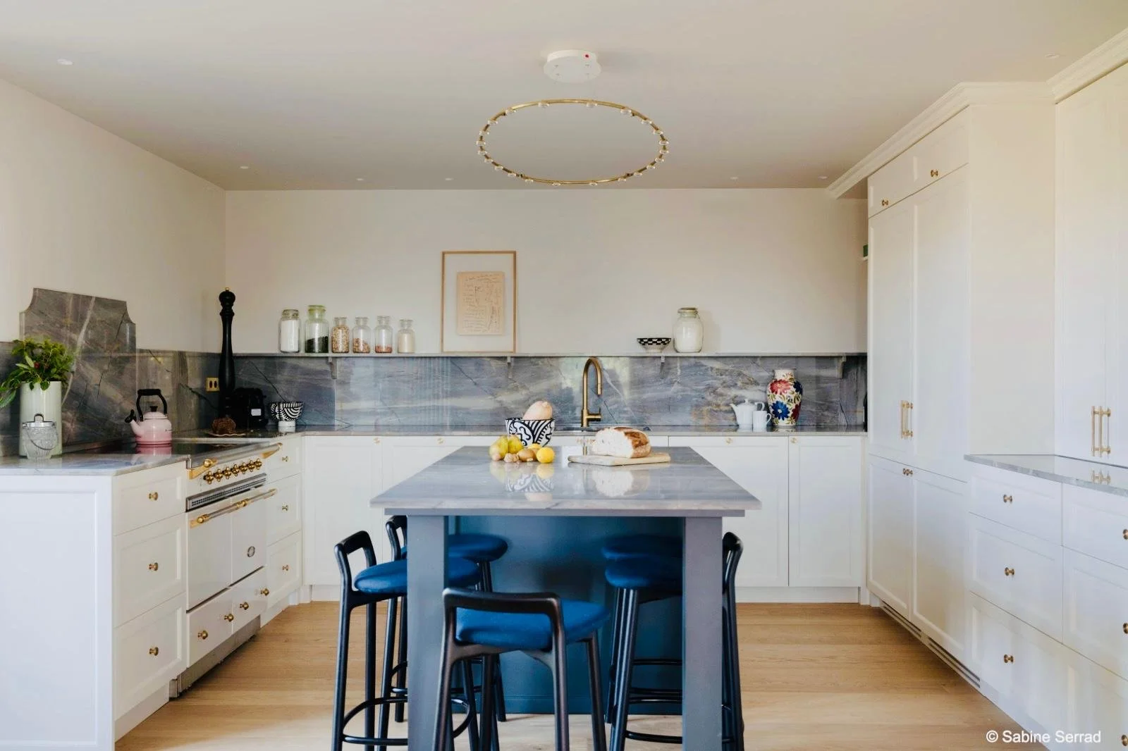 Cuisine moderne avec îlot central, comptoir en marbre, armoires blanches avec poignées en or, lavabo au centre, étagère avec bocaux en verre et vaisselle, décoration minimaliste, mobilier en bois et accents dorés.