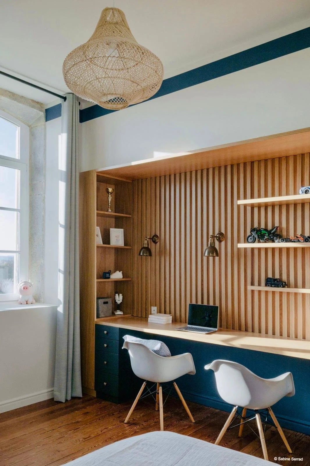 Grand bureau de l’Orangerie Lyon avec étagères en bois intégrées, objets décoratifs et jouets de voitures, espace de travail avec ordinateur portable et deux chaises modernes blanches, lampes murales et suspension en rotin, grande fenêtre avec rideau