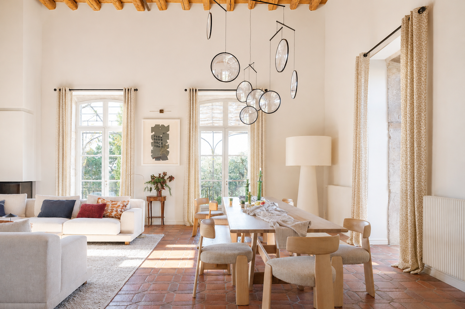 Salon lumineux de l’Orangerie à Lyon avec deux canapés blancs, grande fenêtre avec rideaux légers, tapis clair, plantes vertes, lampadaire sur pied et plafond à poutres en bois, ambiance moderne et élégante pour shootings et tournages.