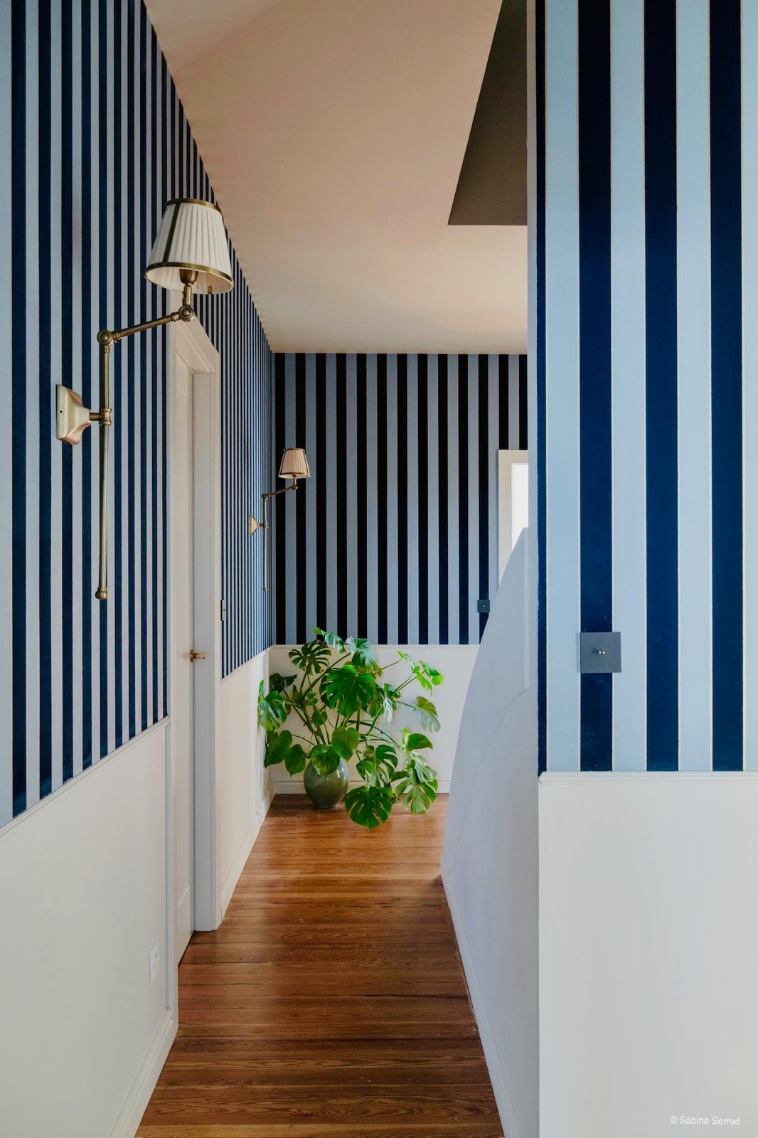 Couloir avec murs à rayures bleues et blanches, lampes murales, planche en bois, plante verte dans un vase, fenêtre en arrière-plan.