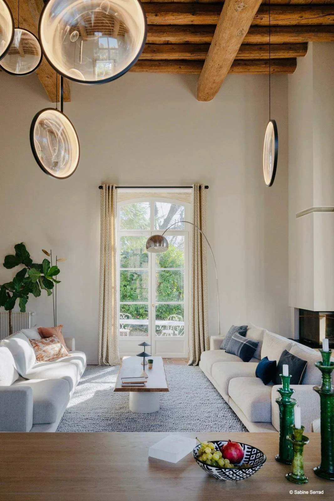 Salon lumineux avec deux canapés blancs, une grande fenêtre avec des rideaux beiges, une lampe arquée, un tapis gris, une cheminée et des décorations vertes, un luminaire suspendu en miroir, et un plateau avec du fruit et des bougeoirs verts.