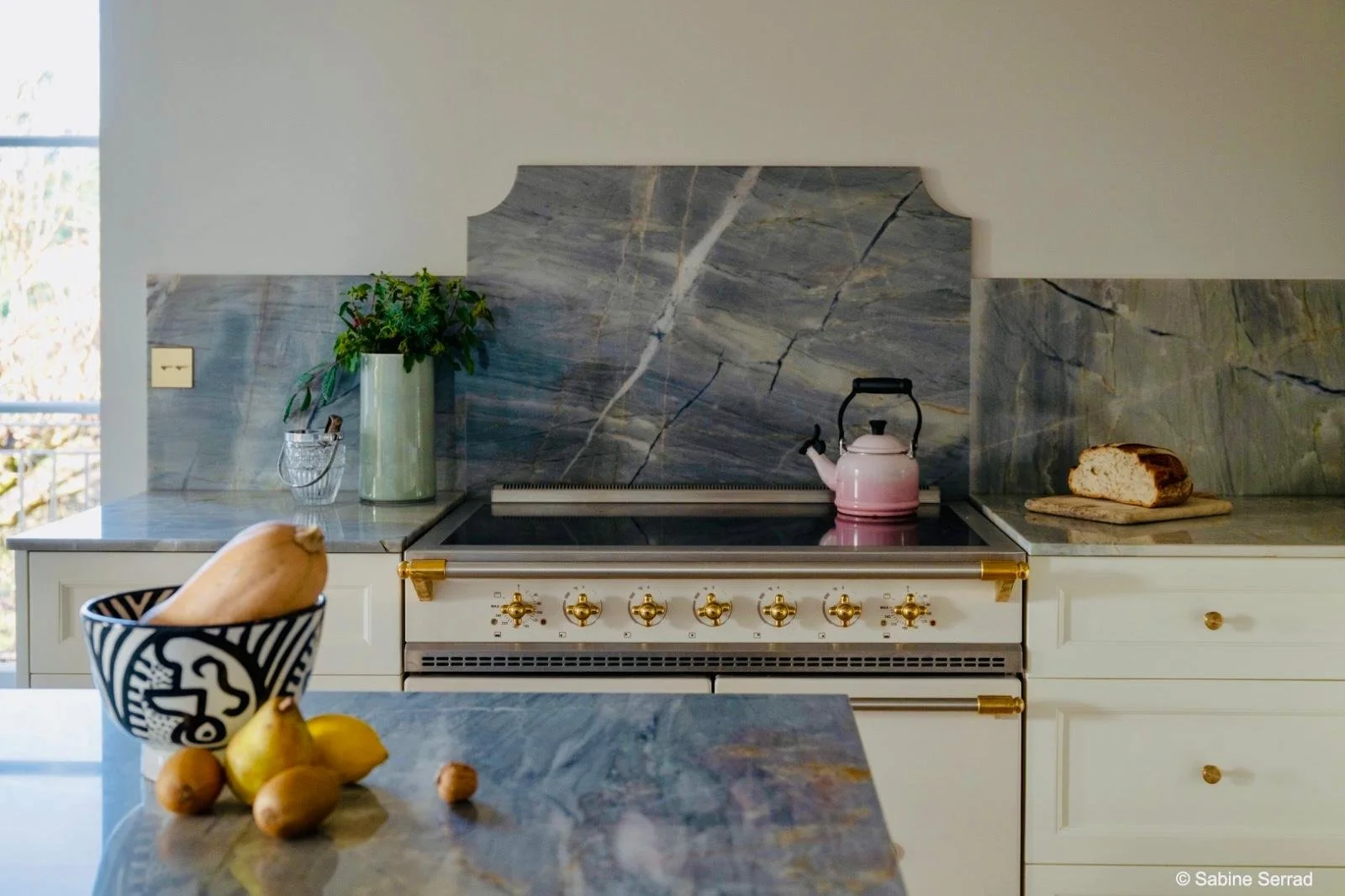Cuisine avec piano blanc, plaque vitrocéramique et plan de travail en marbre gris, théière rose, pain sur une planche, vase avec des plantes, bol avec fruits, et un fond de fenêtre ensoleillée.