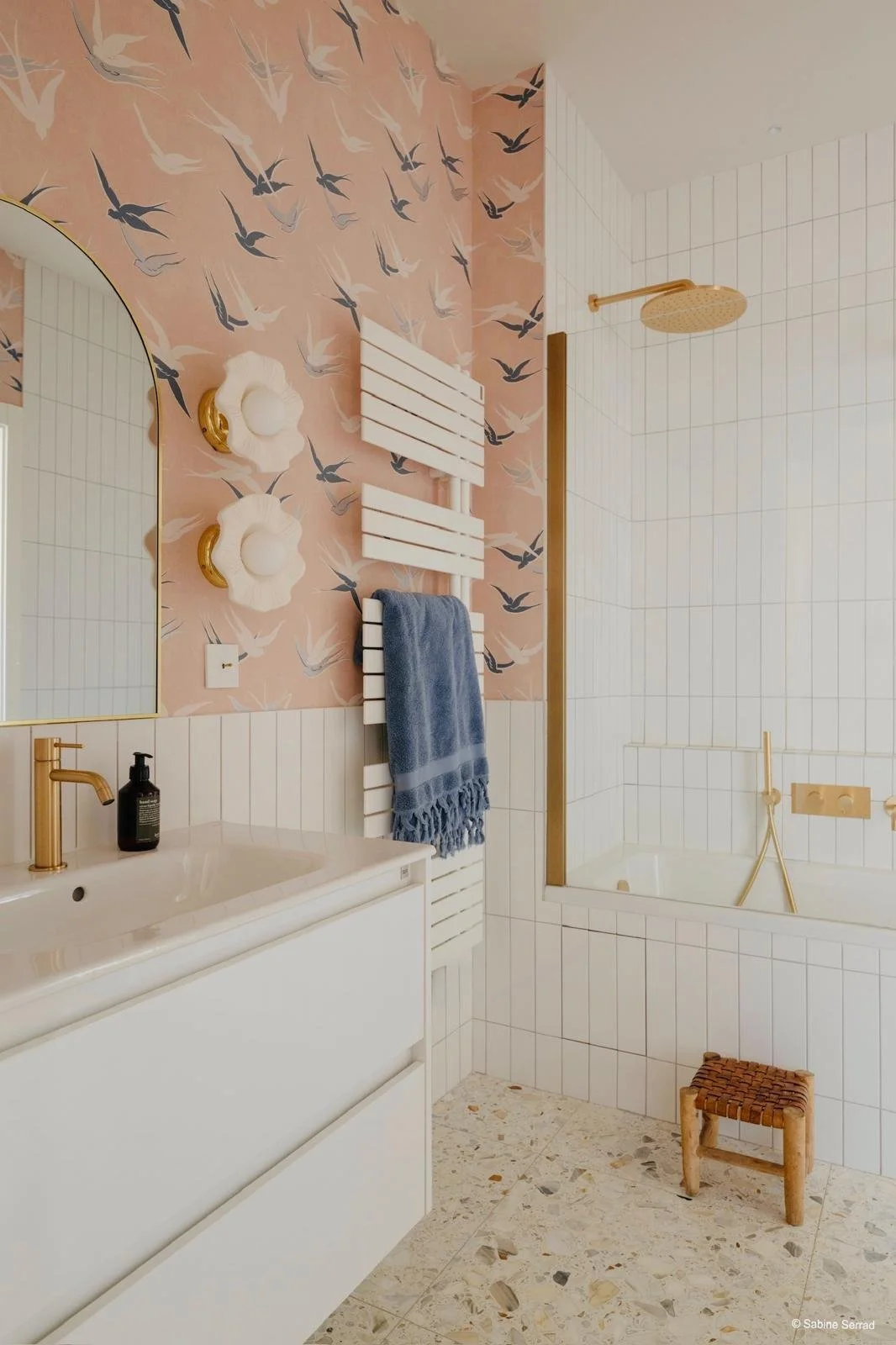 Salle de bain moderne avec murs en carrelage blanc et papier peint avec motif de mouettes, grande miroir rond, lavabo blanc avec robinet en or, sèche-serviettes blanc, serviette bleue, douche avec plafond en or, baignoire encastrée, petit tabouret en