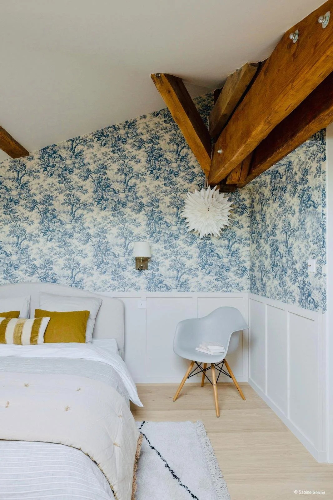 Une chambre avec un mur tapissé d'un papier peint bleu et blanc à motif de nature, un lit avec des coussins marron et blanc, une lampe murale et un fauteuil blanc avec des livres posés dessus.