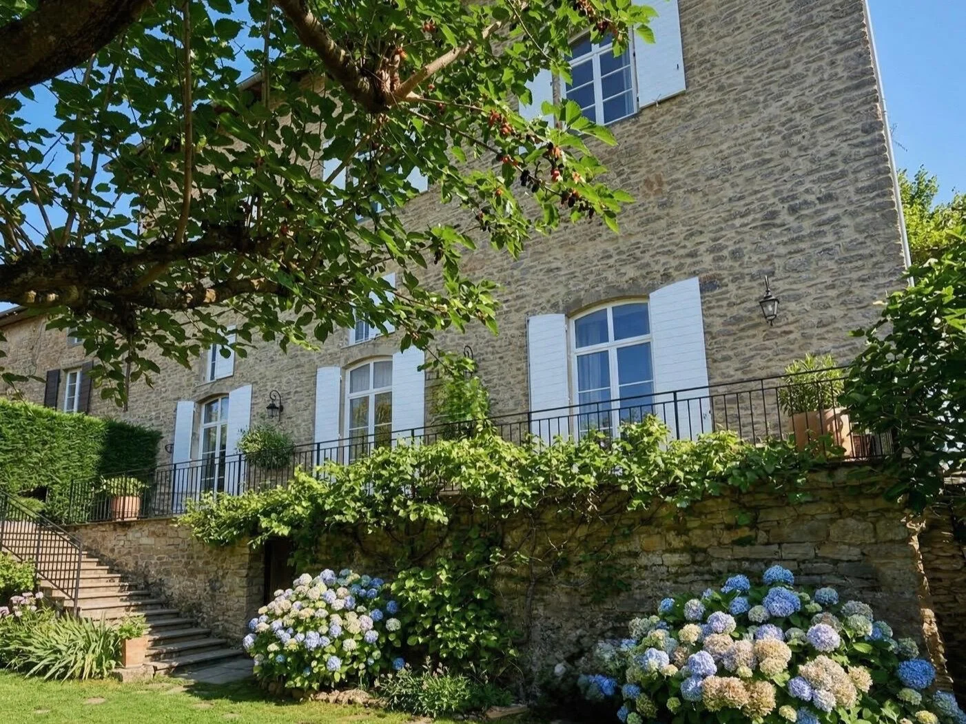 Maison en pierre avec fenêtres à volets blancs, balcon avec des plantes en pot, escaliers en pierre, hortensias en fleurs dans le jardin, arbres verts, ciel bleu.