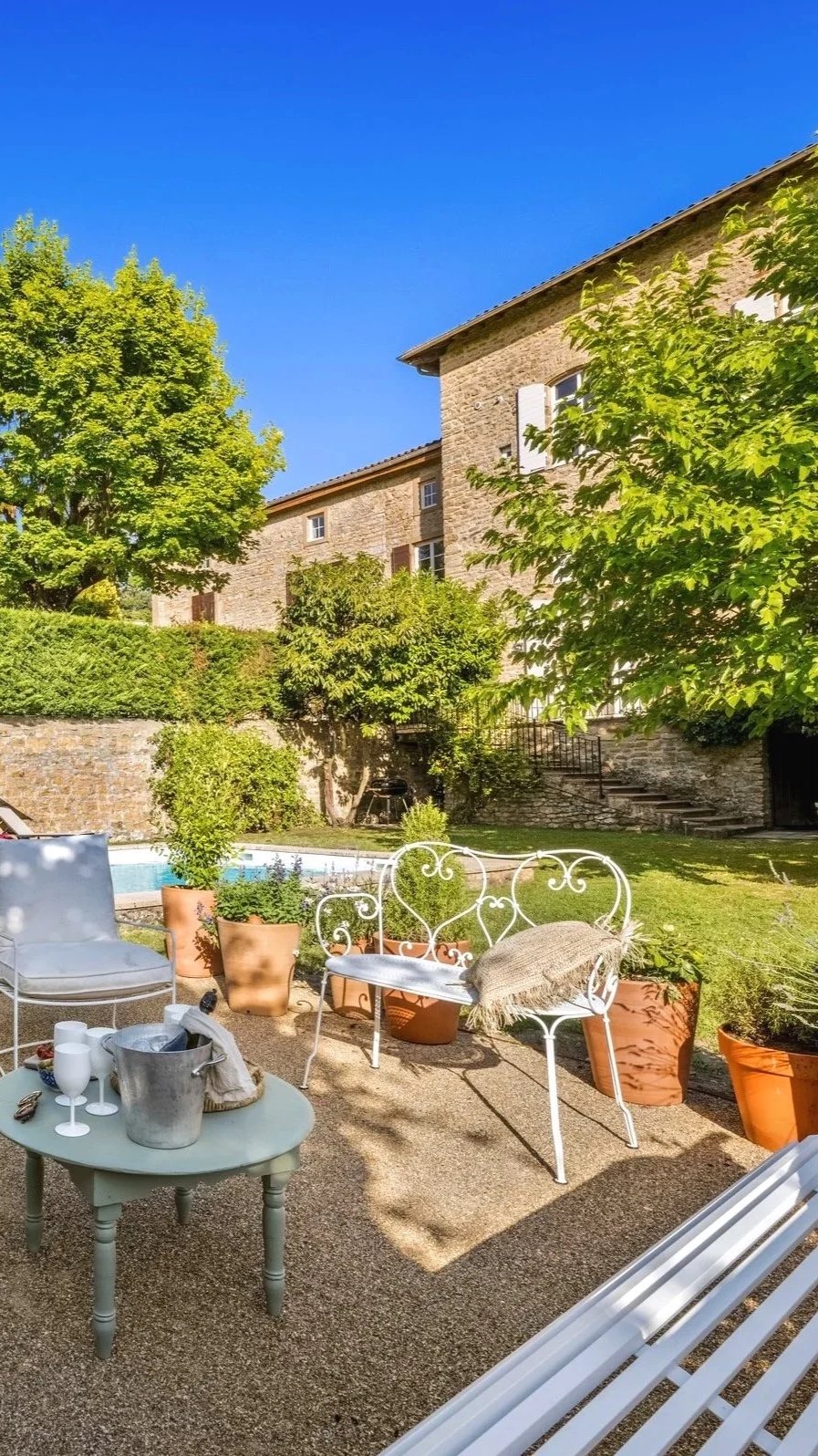 Jardin extérieur avec bancs en fer blanc, table, pots de fleurs, arbres verdoyants et maison en pierre sous un ciel bleu.