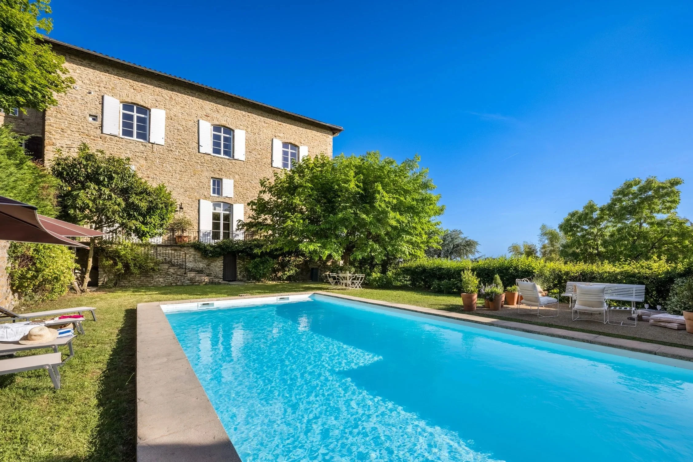 Maison en pierre entourée de végétation avec piscine bleue et mobilier d’extérieur dans un jardin, sous un ciel bleu clair, située à l’Orangerie Lyon, idéale pour tournages, shootings photo et productions audiovisuelles.