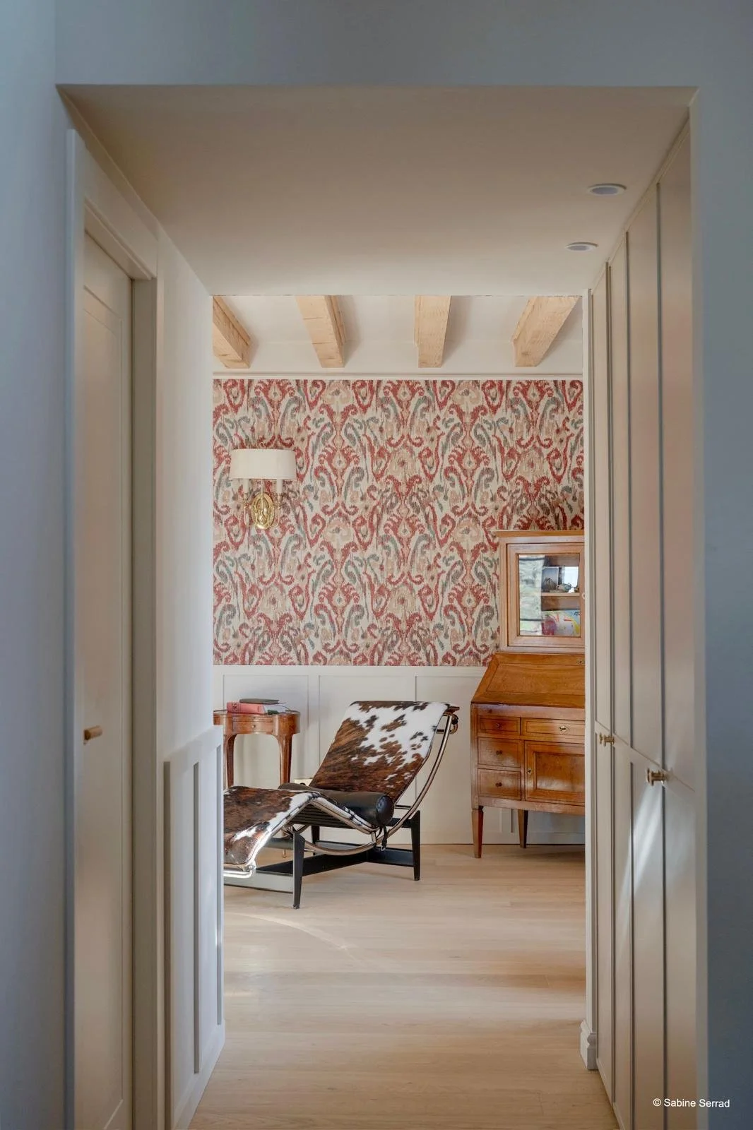 Une salle de séjour avec un mur à papier peint rouge et beige, un fauteuil en cuir avec motif animal, une petite table en bois, une armoire ancienne en bois et un éclairage mural.