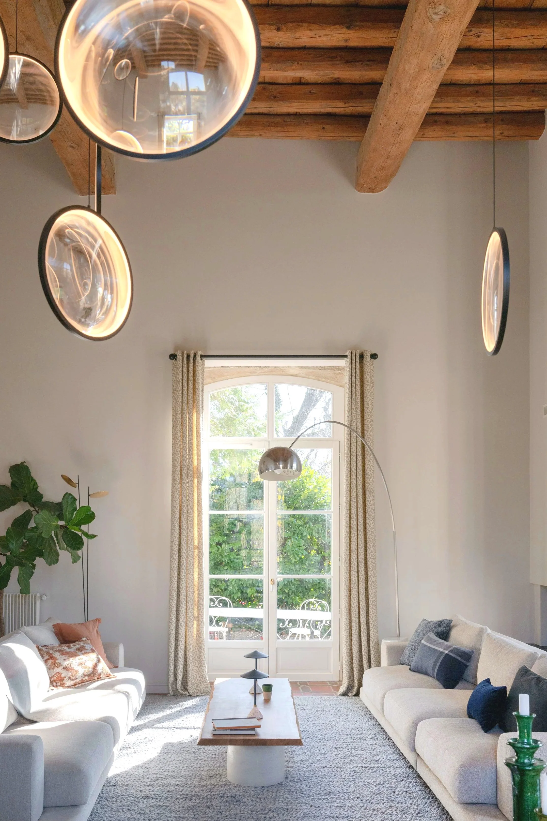 Salon lumineux avec canapés blancs, coussins colorés, un tapis clair, une grande fenêtre avec rideaux, un luminaire suspendu en forme d'arc, un mur en bois au plafond, plantes décoratives et table basse en bois.