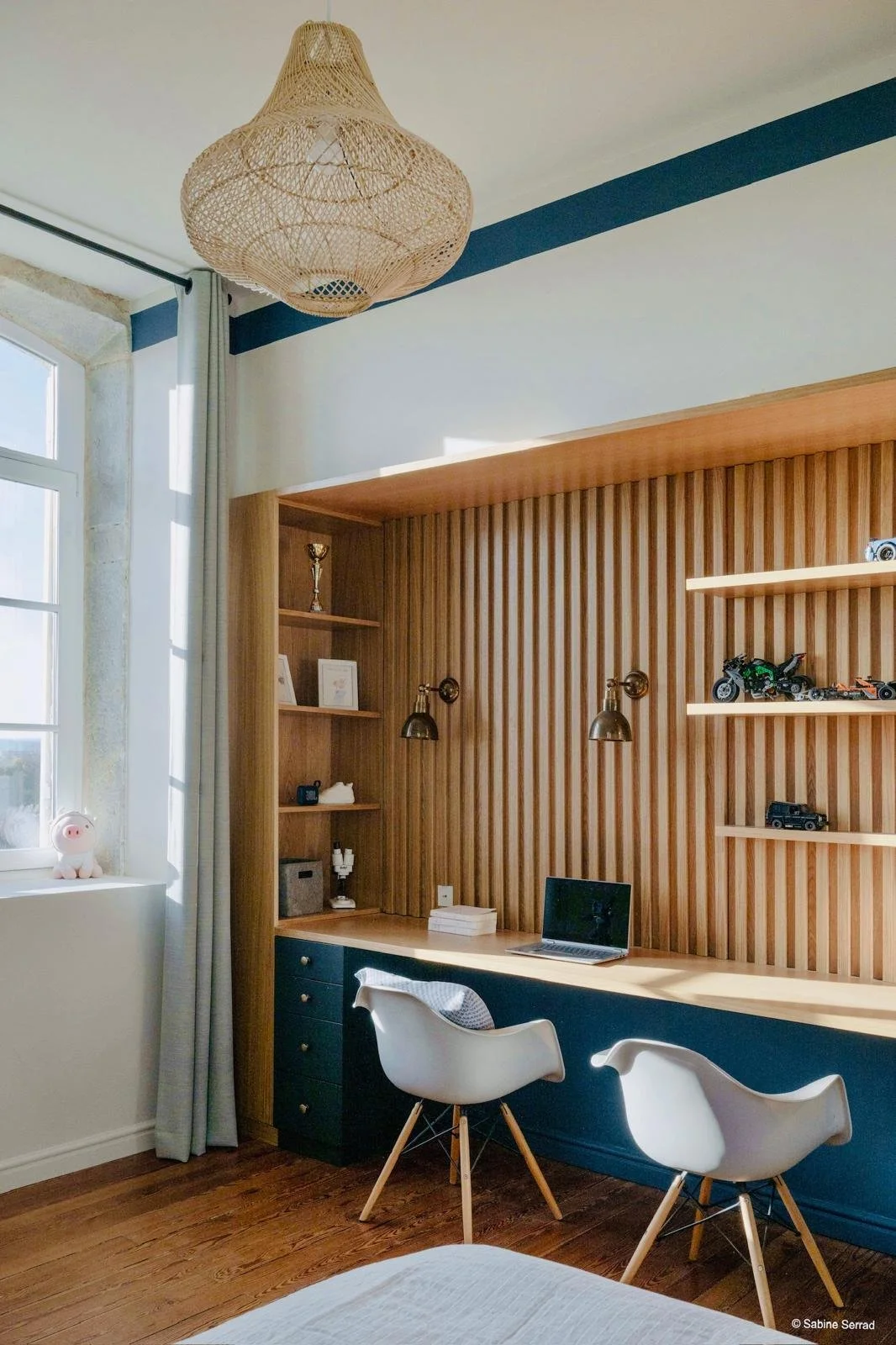 Coin bureau avec une baie vitrée, étagères en bois avec des jouets de voitures, un ordinateur portable, deux chaises modernes, une lampe murale, un plafond avec un lampe en rotin et une fenêtre avec un rideau blanc.