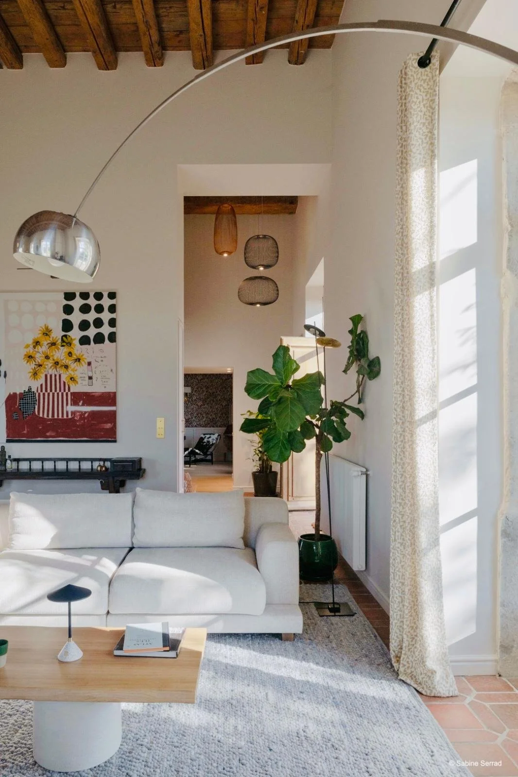 Salon intérieur lumineux de l’Orangerie Lyon avec canapés beiges, table en bois et chaises assorties dans une ambiance épurée et contemporaine. Grandes fenêtres avec rideaux laissant entrer une lumière naturelle abondante, lampes modernes, poutres ap