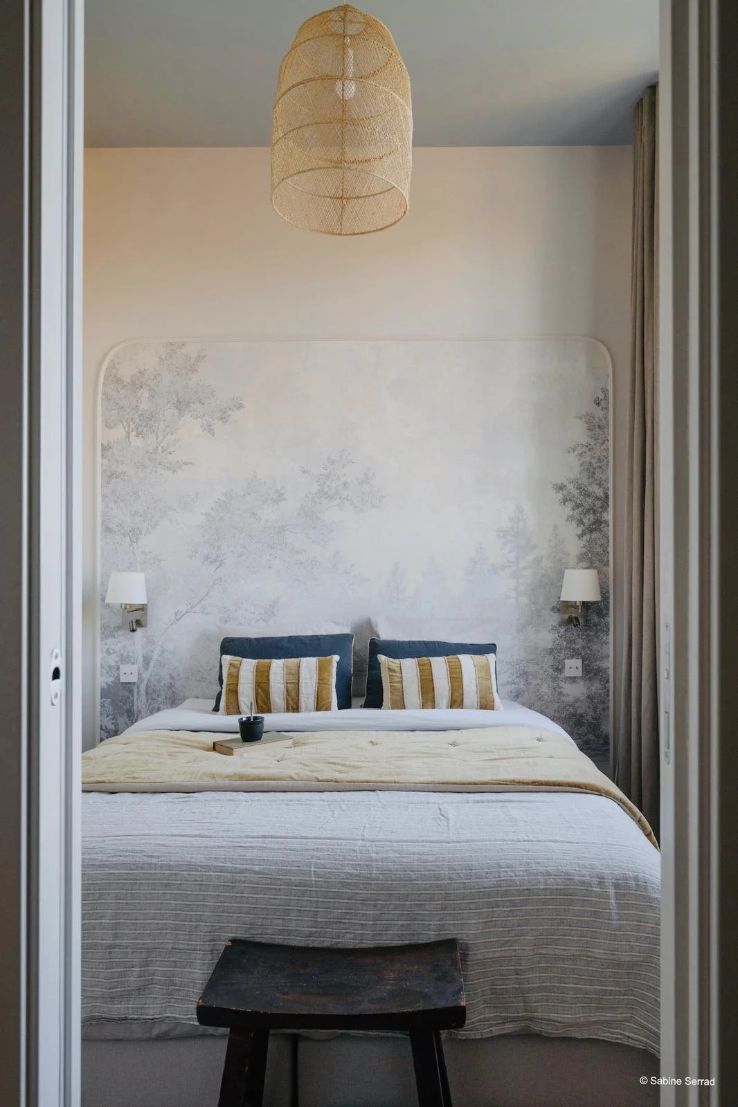 Chambre avec un lit double, deux coussins à rayures, lampes murales, peinture murale de forêt, lampadaire en rotin suspendu, rideaux gris, petit tabouret en bois au premier plan.