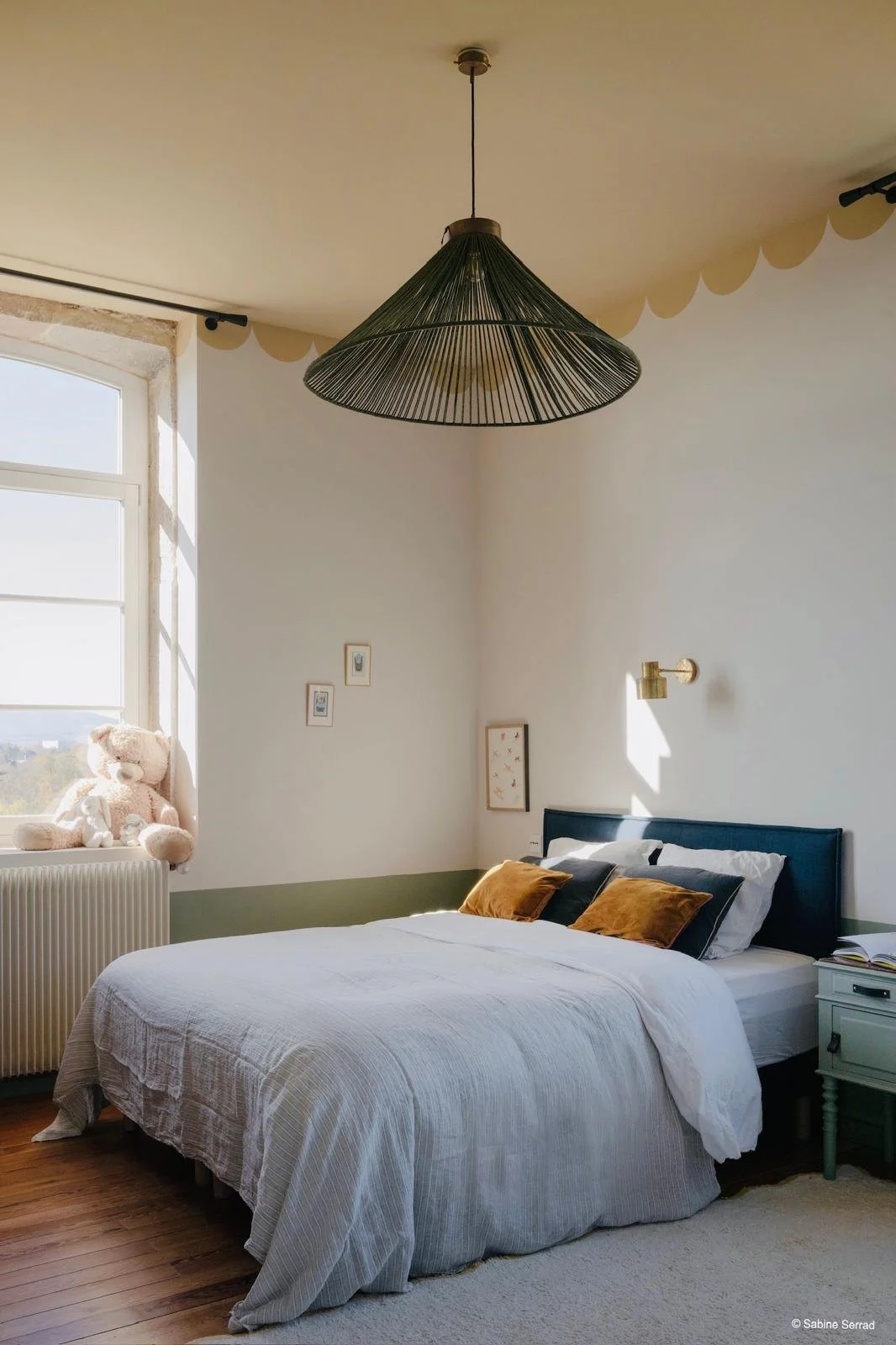 Chambre avec un lit double, oreillers marrons et noirs, une tête de lit bleue, une lampe murale, une étagère blanche, une fenêtre avec une poupée ou ours en peluche sur le rebord, et des petits tableaux.