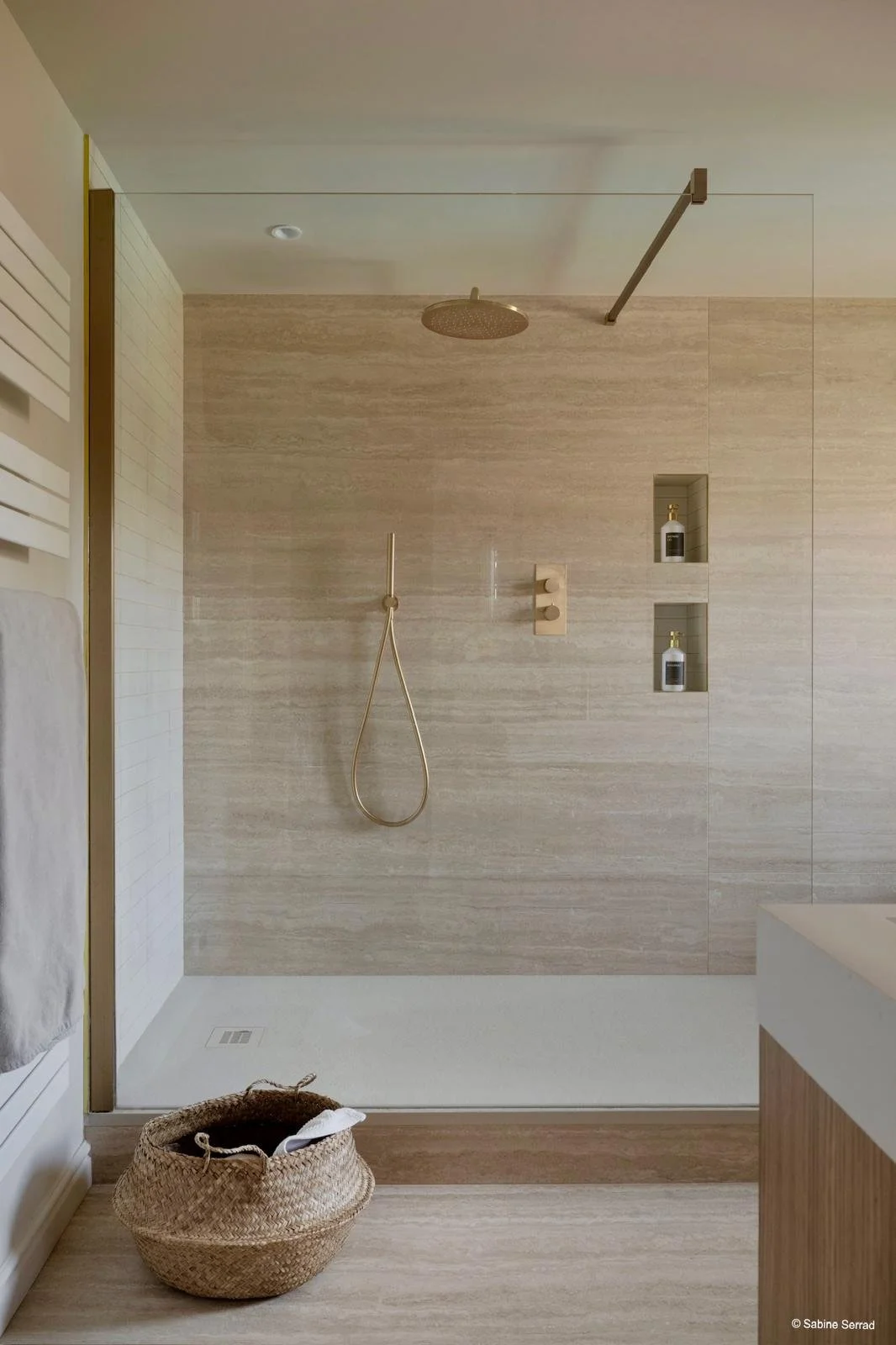 Douche moderne avec douchette ronde au plafond, murs en pierre beige, niche avec produits de douche, panier en osier avec linge, sol beige, partie d'une salle de bain minimaliste.