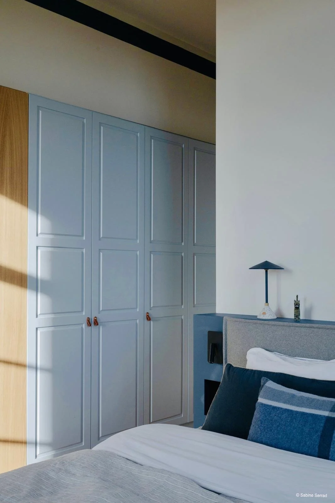 Chambre avec une grande armoire bleue à portes moulurées, un lit avec des coussins noirs et gris, une table de chevet avec une lampe et une décoration.