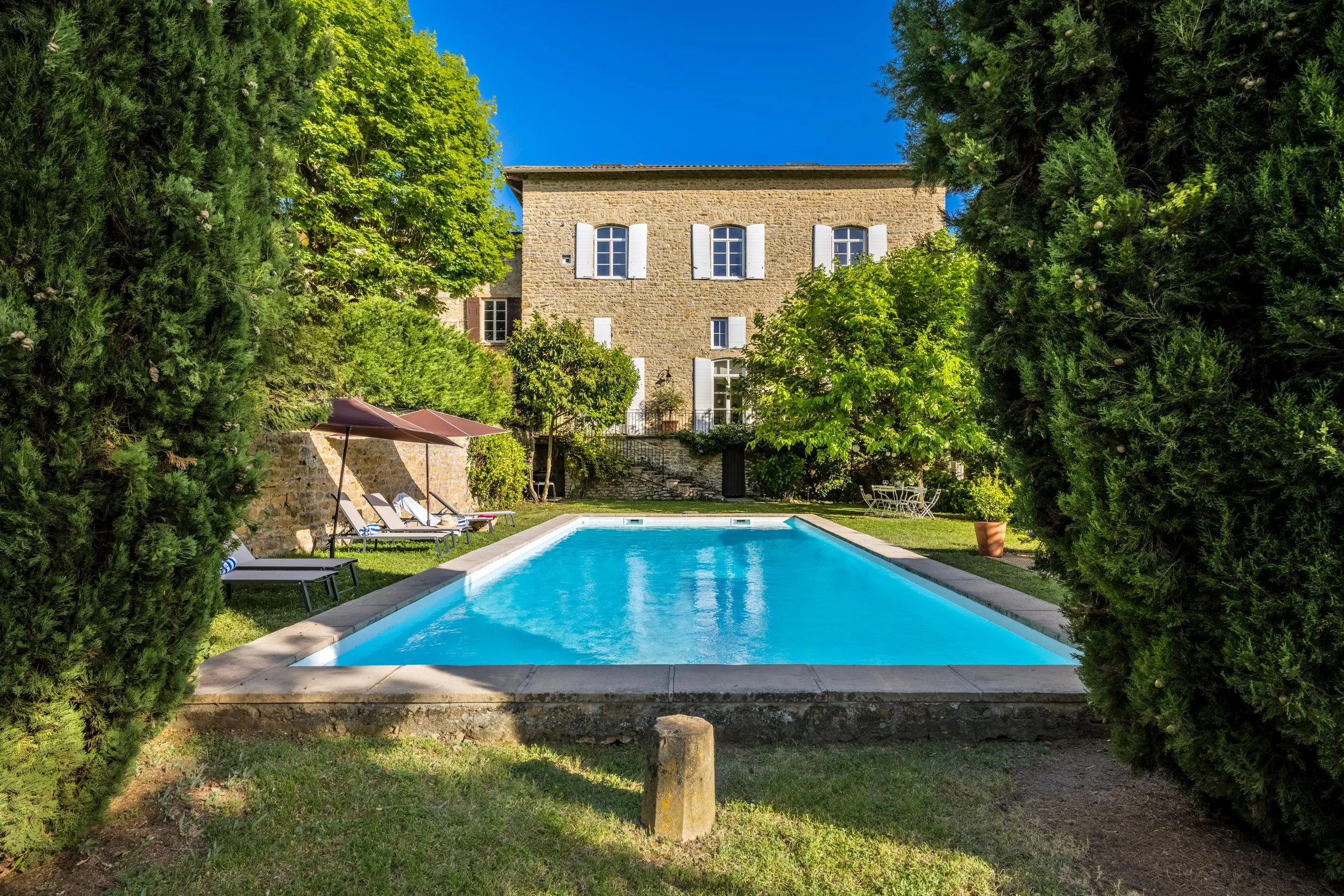 maison en pierre avec fenêtres à volets blancs, piscine entourée de chaises longues, arbres verts, ciel clair