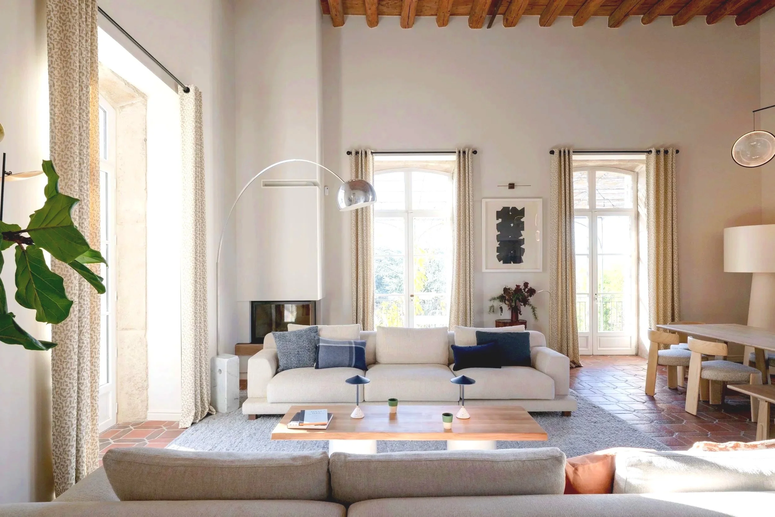 Un salon lumineux avec des grandes fenêtres, un canapé beige avec des coussins gris et bleus, une table basse en bois, et des plantes vertes, décoré dans un style moderne et épuré.