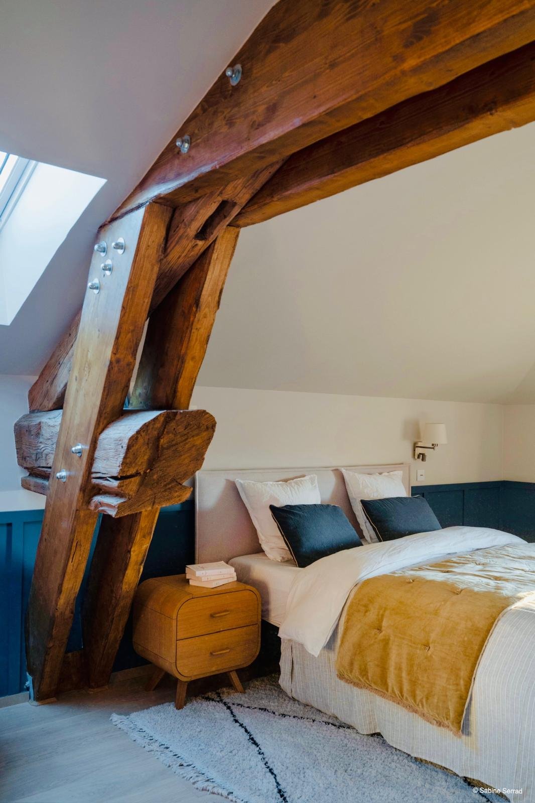 Chambre avec lit, oreillers blancs et noirs, lampadaire, meuble de chevet en bois, poutre en bois apparente au plafond, mur blanc et bleu marine.