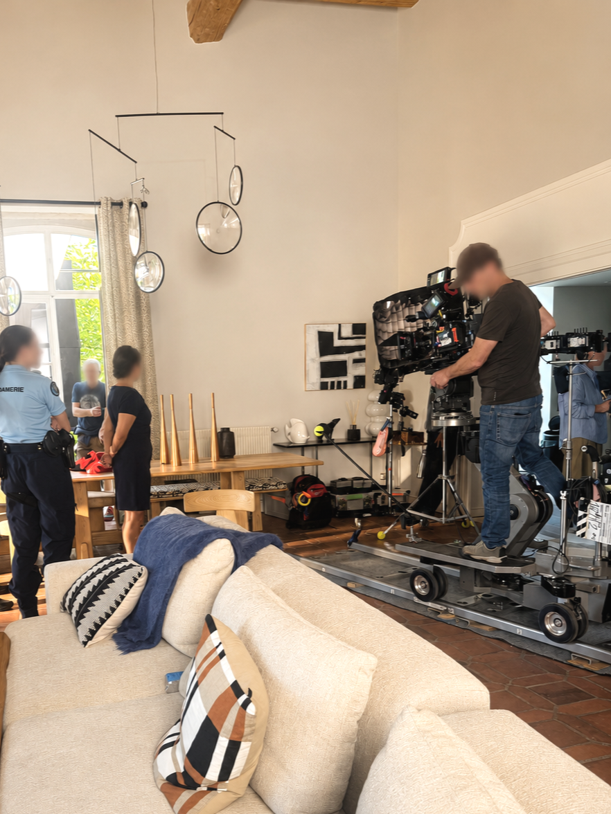 Une scène de tournage dans un salon avec un caméraman sur un steadicam. Quelques personnes discutent, dont une police. Le salon est meublé avec un canapé beige, des coussins à motifs et une table en bois. Il y a des décorations modernes et une grande fenêtre laissant entrer de la lumière naturelle.