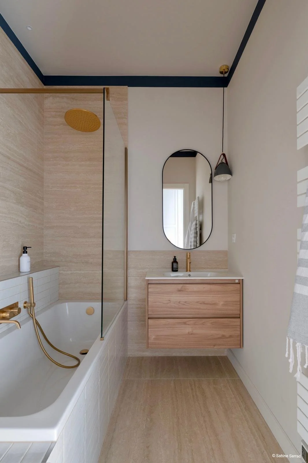 Salle de bain moderne avec baignoire blanche, robinetterie en or, cabine de douche avec paroi en verre, vasque sur meuble en bois clair, miroir ovale, lampadaire suspendu, murs en beige et accents en bleu foncé, fenêtre avec volets blancs.