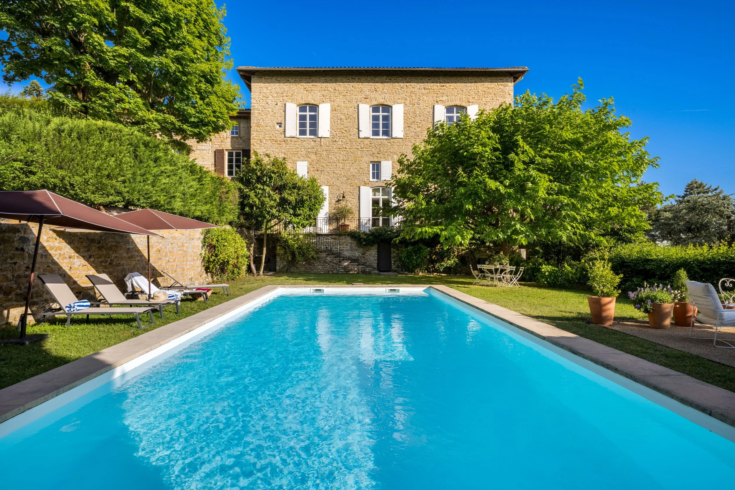 Une maison en pierre avec des volets blancs, entourée d'arbres verts, avec une piscine extérieure entourée de transats, un parasol et des pots de fleurs en terracotta, par une journée ensoleillée avec un ciel bleu clair.