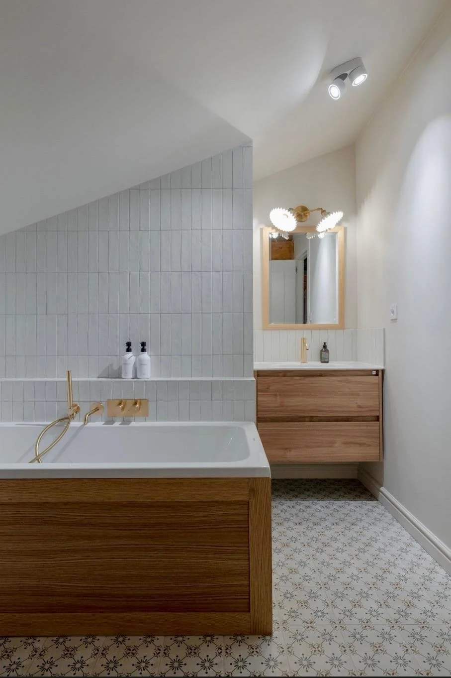 Salle de bain avec une baignoire en bois, un miroir, un lavabo avec un meuble en bois, deux bouteilles de savon sur la baignoire, un éclairage au plafond, et un sol à motifs.