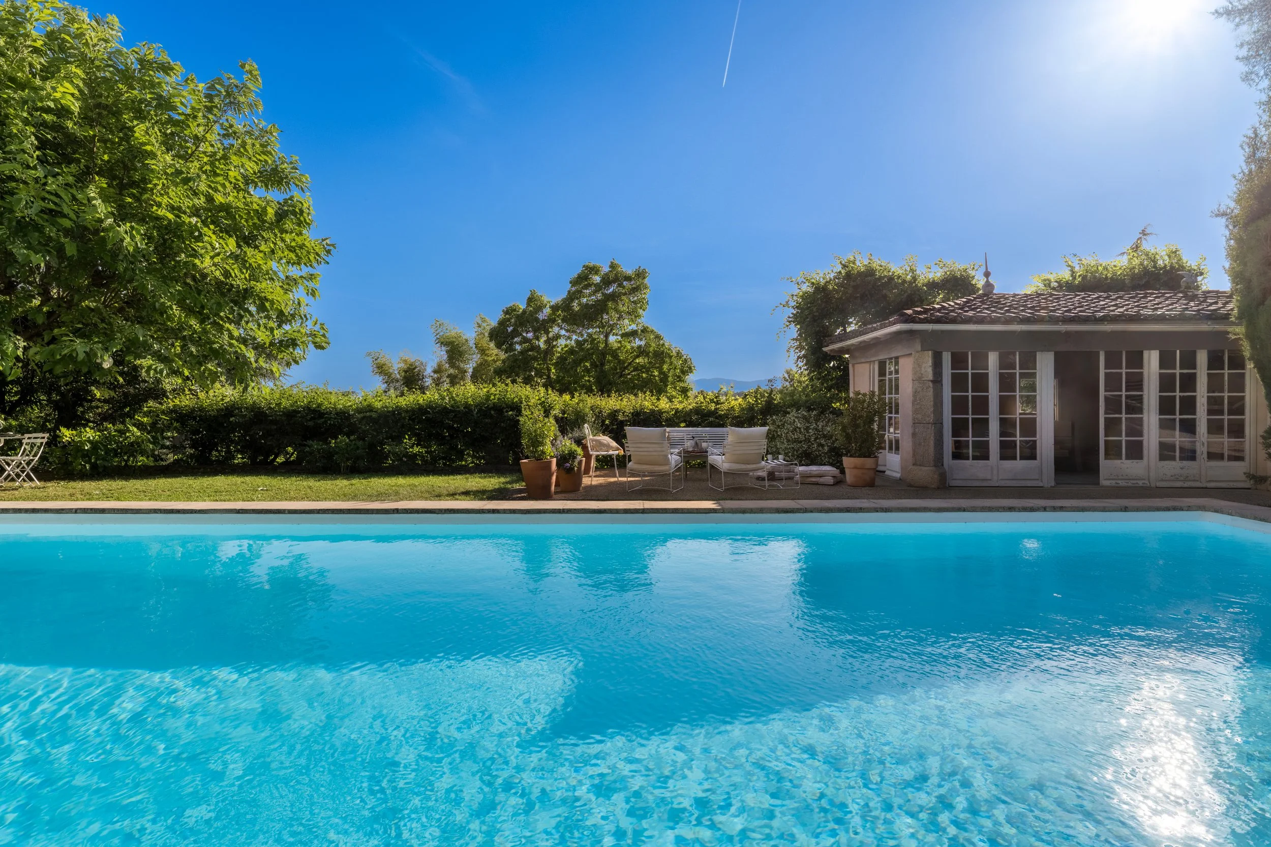 Maison en pierre de l’Orangerie Lyon avec volets blancs, entourée d’arbres verts et baignée de lumière naturelle, avec piscine extérieure, transats, parasol et pots de fleurs en terre cuite dans un jardin verdoyant sous ciel bleu clair, idéale pour t