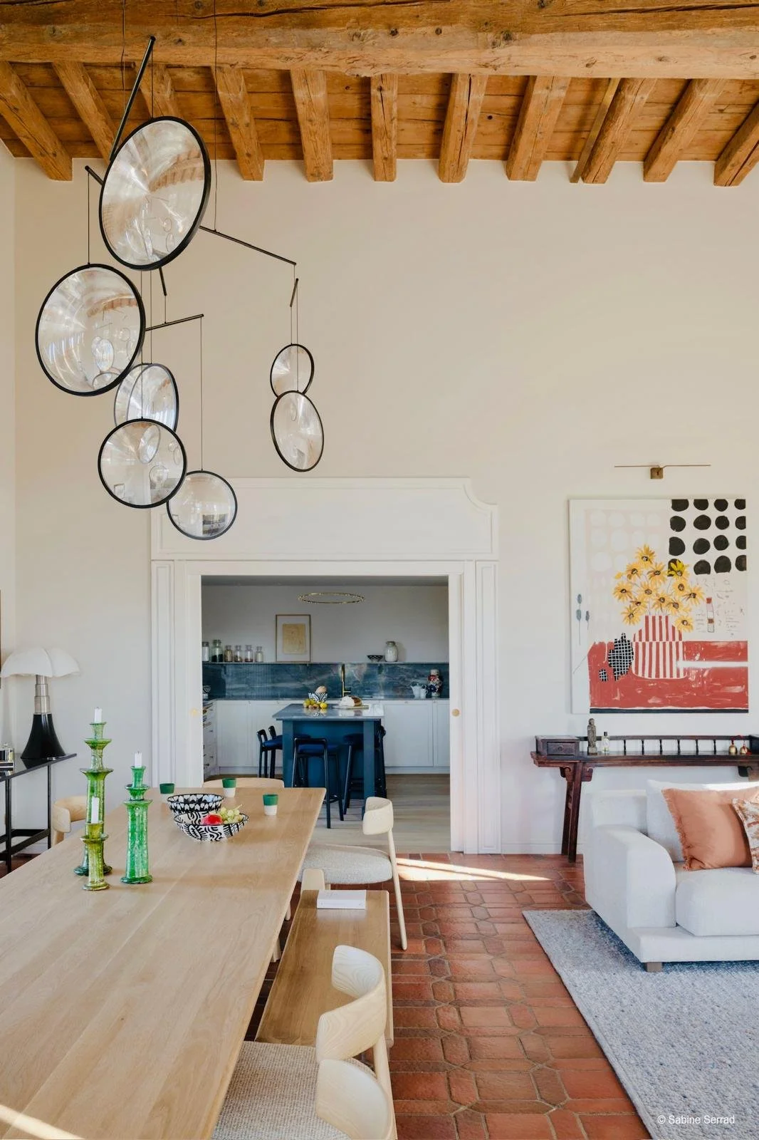 Salon et cuisine modernes avec plafond en bois, lampe suspendue, table en bois avec chaises, décoration colorée, canapé blanc avec coussins, oeuvres d'art sur les murs, sol en carreaux de terre cuite.