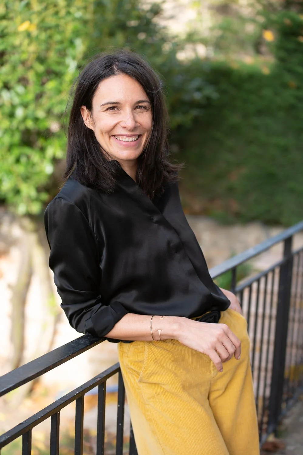 Une femme sourit, portant une chemise noire en satin et un pantalon jaune, posant contre une rampe en extérieur avec un arrière-plan verdoyant.