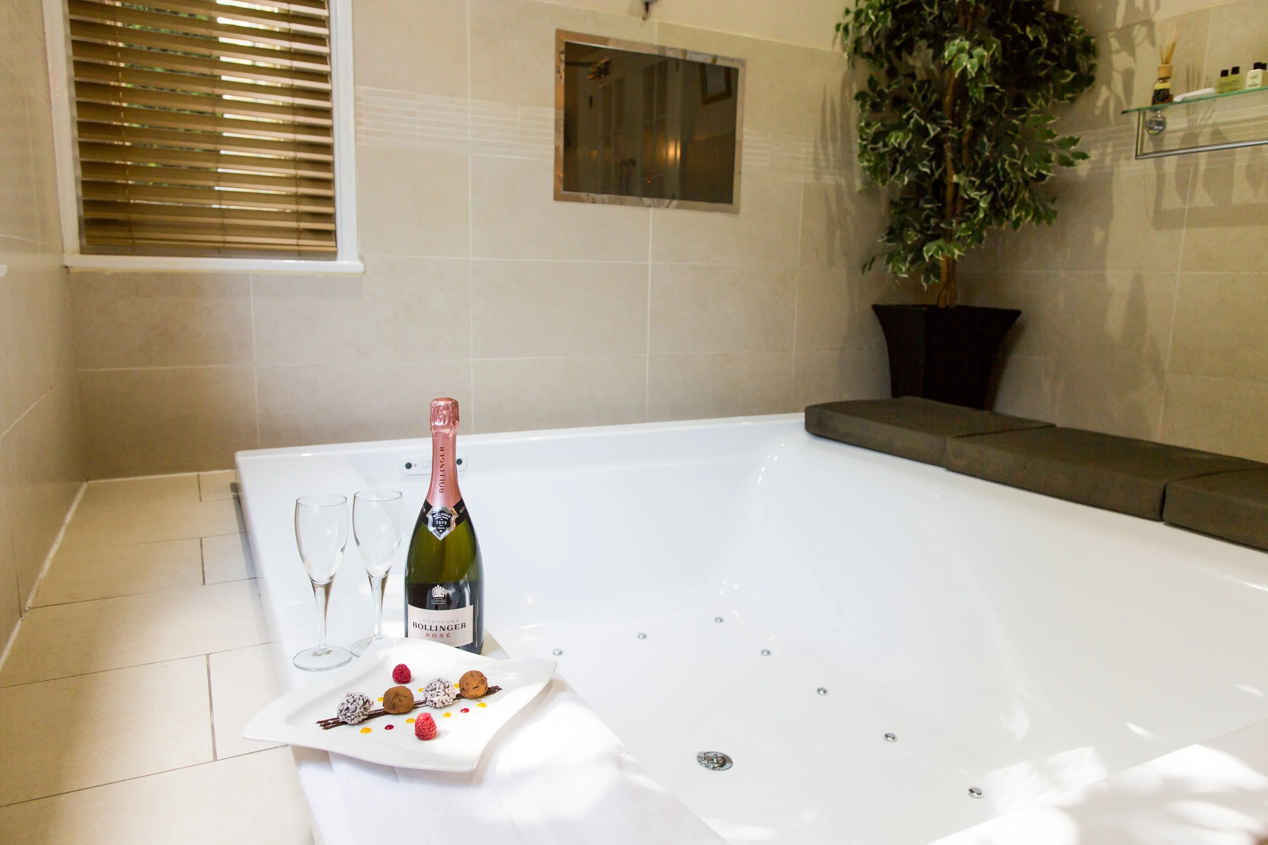 A luxurious bathroom with a large white jacuzzi tub, a bottle of pink sparkling wine, two empty champagne glasses, a plate with assorted chocolates and raspberries, beige tiled walls and floor, a small window with blinds, a mirror, a potted plant, an