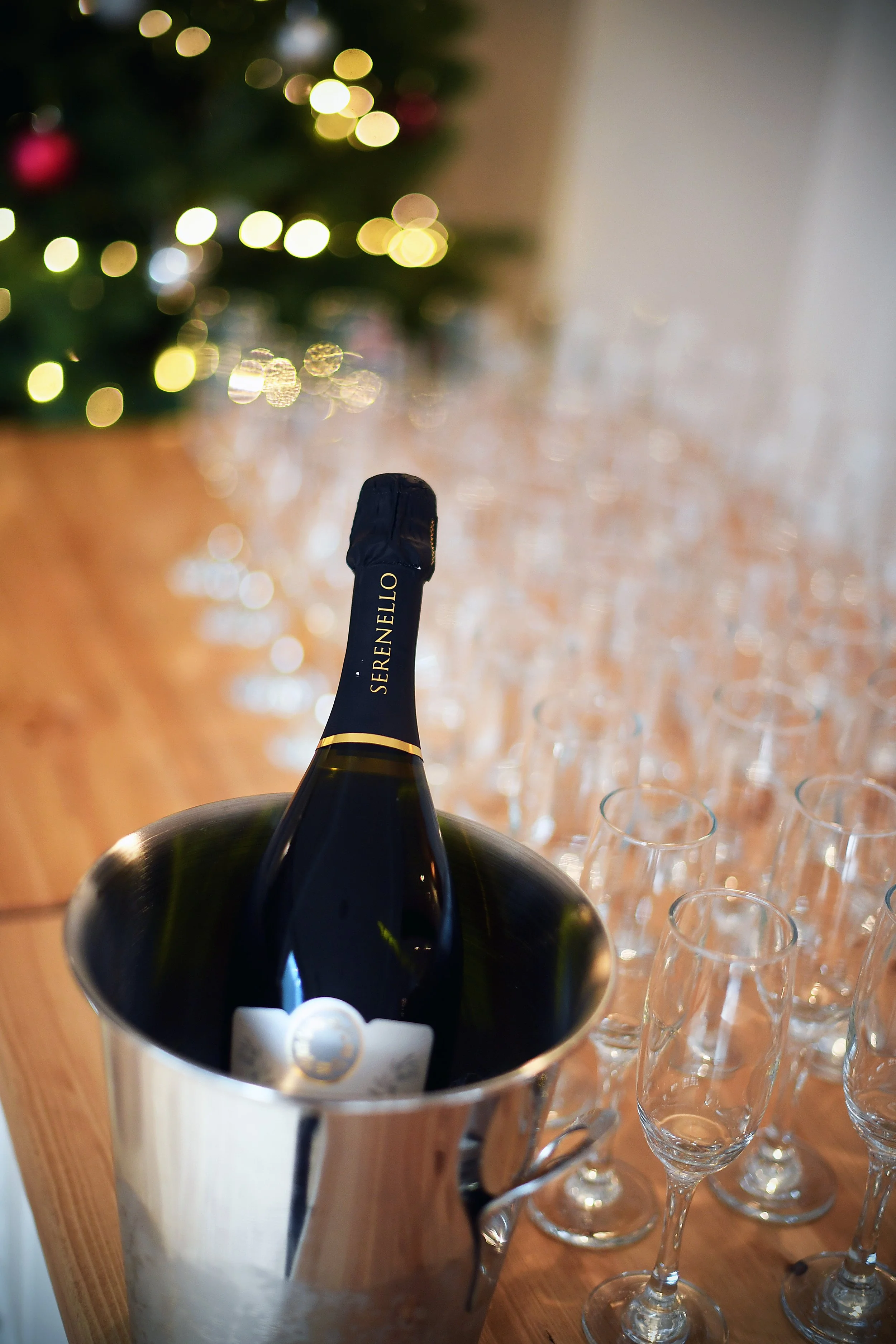 A bottle of sparkling wine in an ice bucket surrounded by empty champagne flutes on a wooden table, with a blurred Christmas tree in the background.