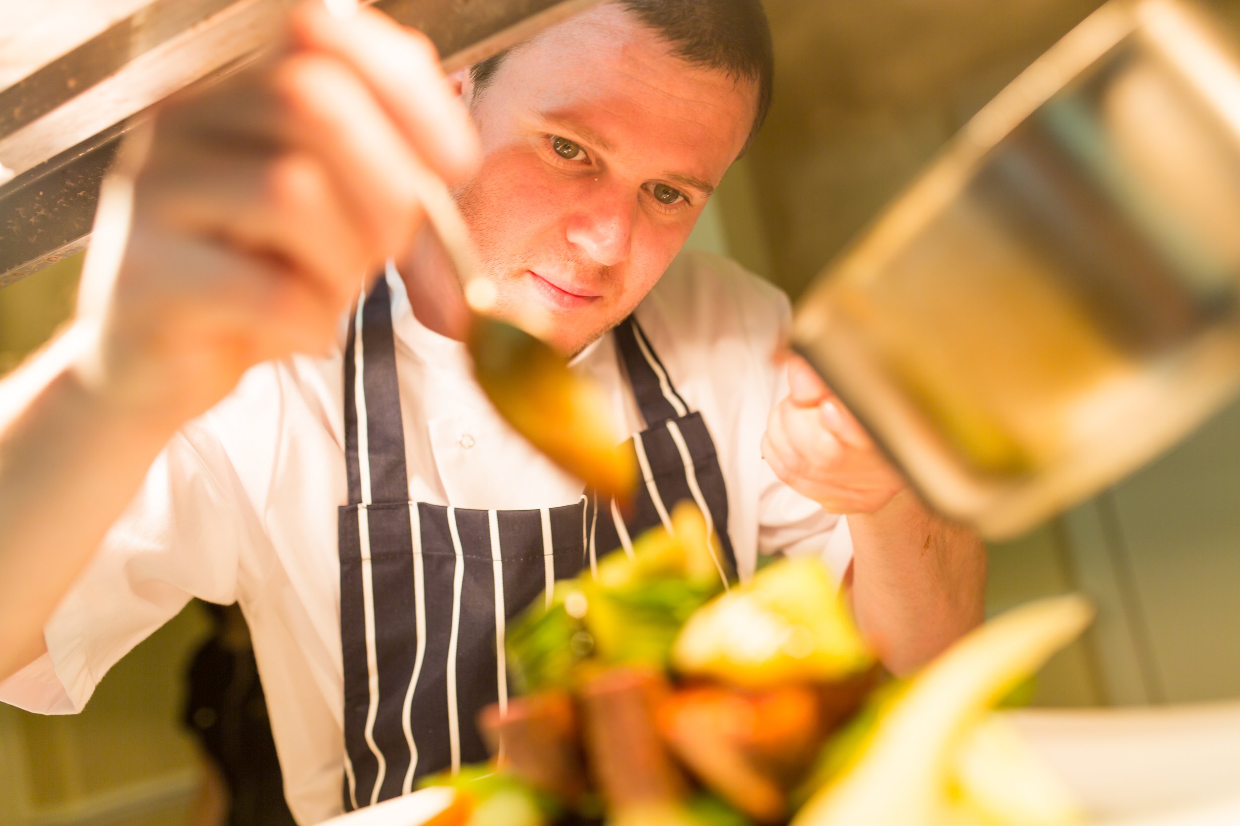 A chef in a striped apron preparing food, seen from below with fresh ingredients in focus.