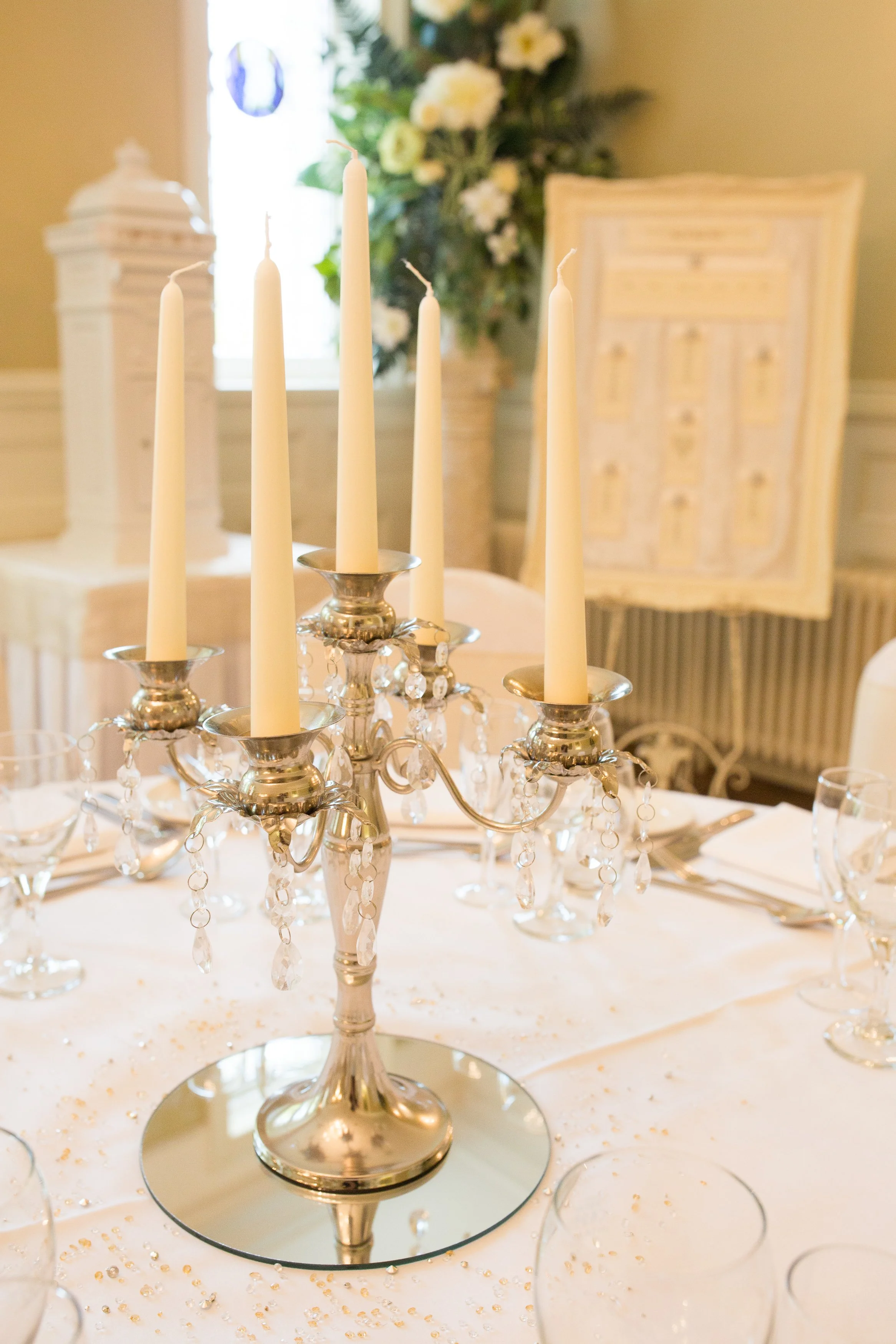 Elegant dining table set with a silver candelabra holding five tall, unlit white candles, surrounded by clear wine glasses and white napkins, in a room with a floral arrangement and a window in the background.