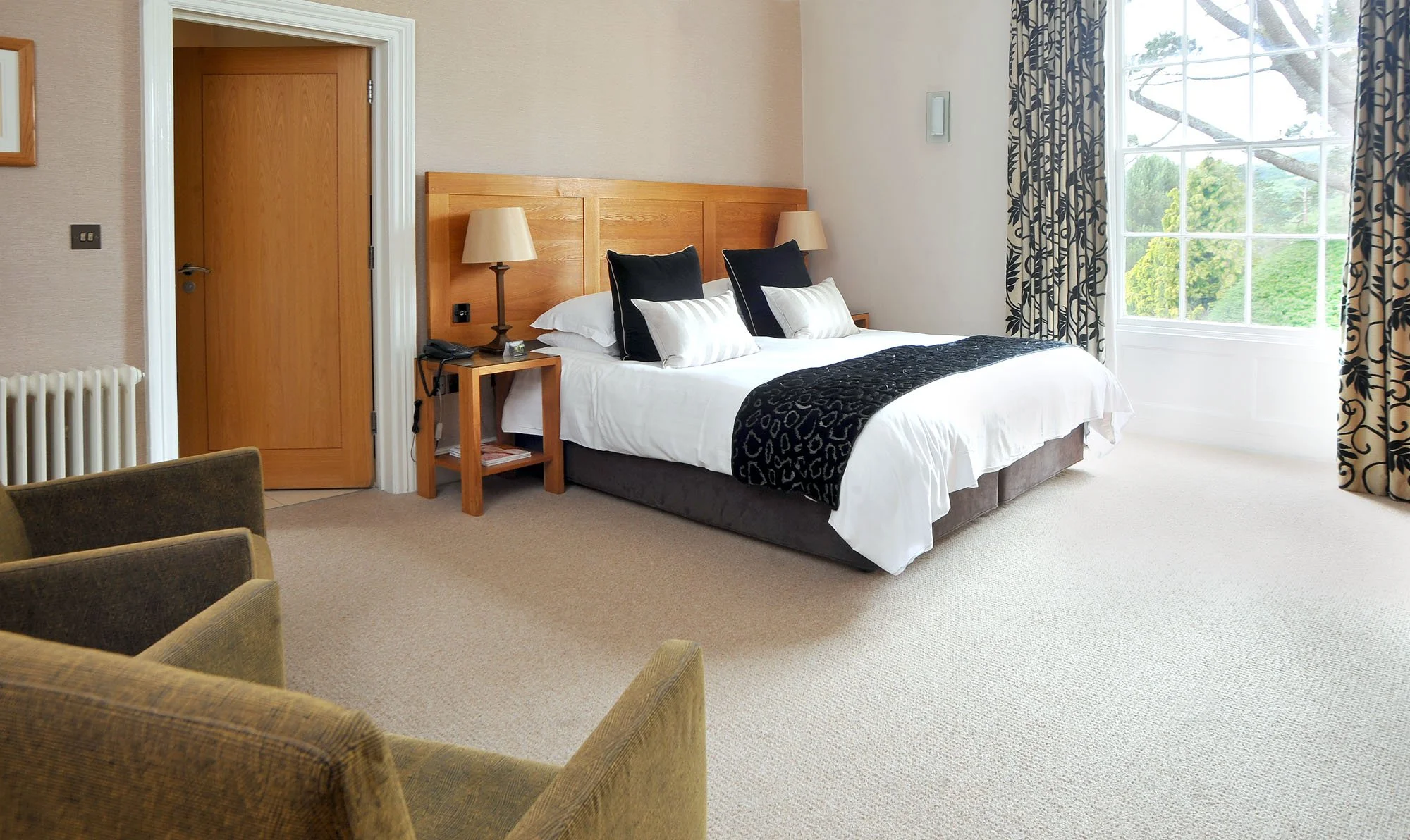 A neatly made hotel room with a large bed, black and white pillows, a wooden headboard, and a window with patterned curtains showing green trees outside.