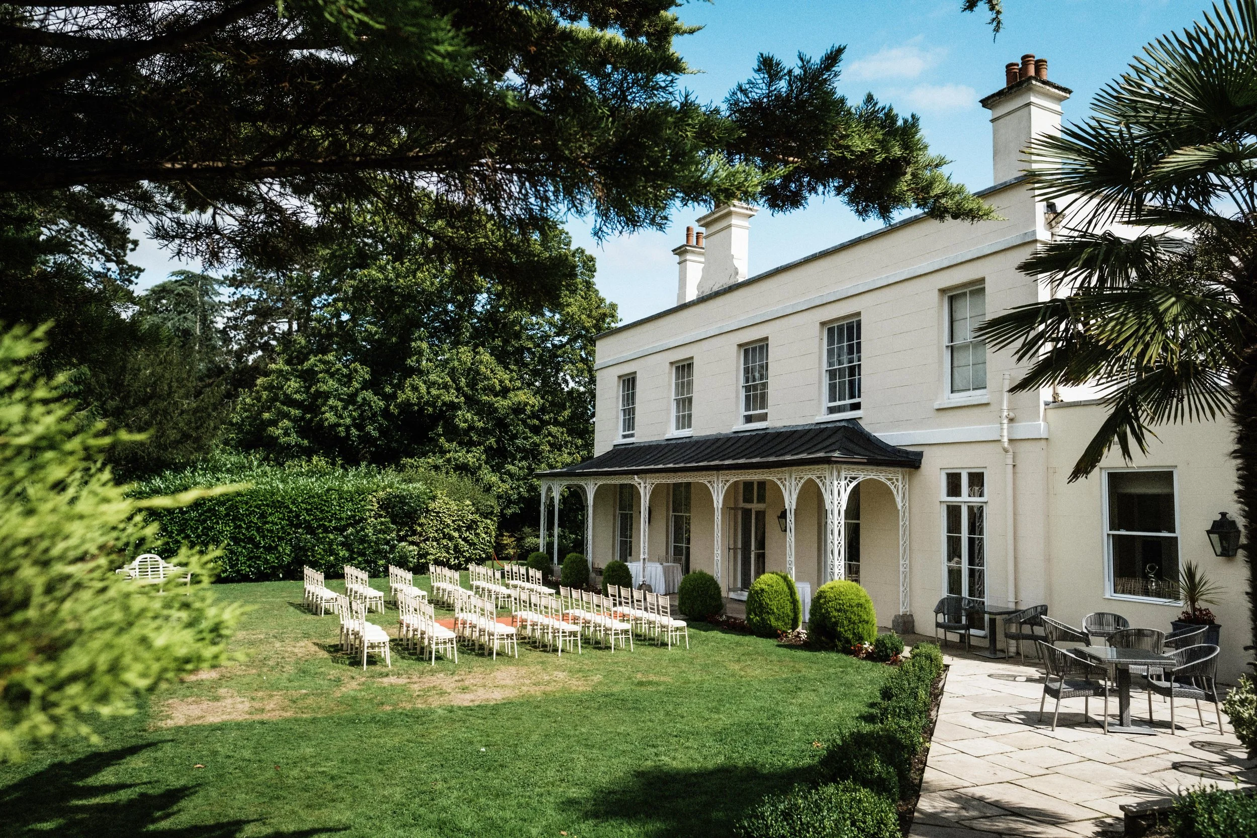 A large white house or manor with a black roof, surrounded by a lush green garden. The garden has white chairs arranged in rows, possibly for an outdoor event, and a patio with a table and chairs. Tall trees and bushes surround the property, with a b
