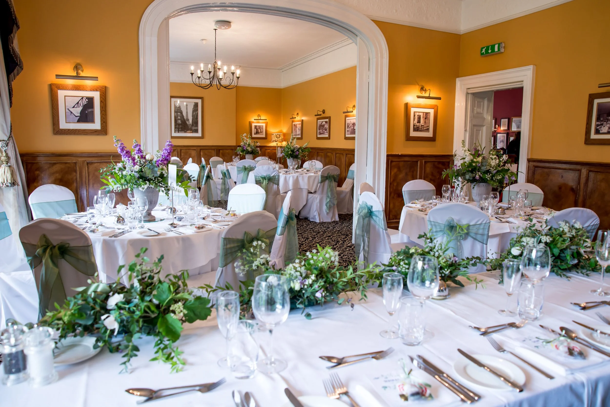 Elegant banquet room decorated with round tables covered with white tablecloths, each with floral centerpieces, glassware, and silverware, set up for a formal event or wedding reception, with yellow walls, framed artwork, and a chandelier.