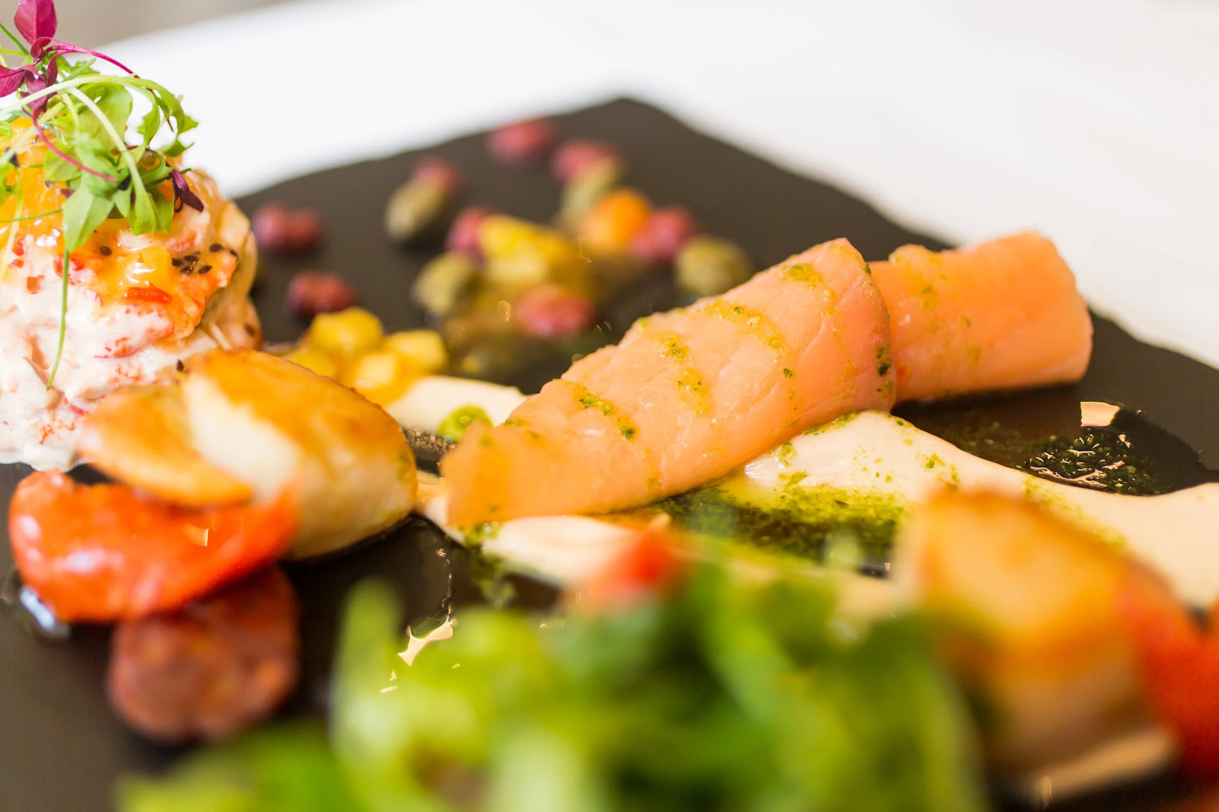 A gourmet dish with cured salmon, creamy sauce, roasted vegetables, and microgreens on a black plate.