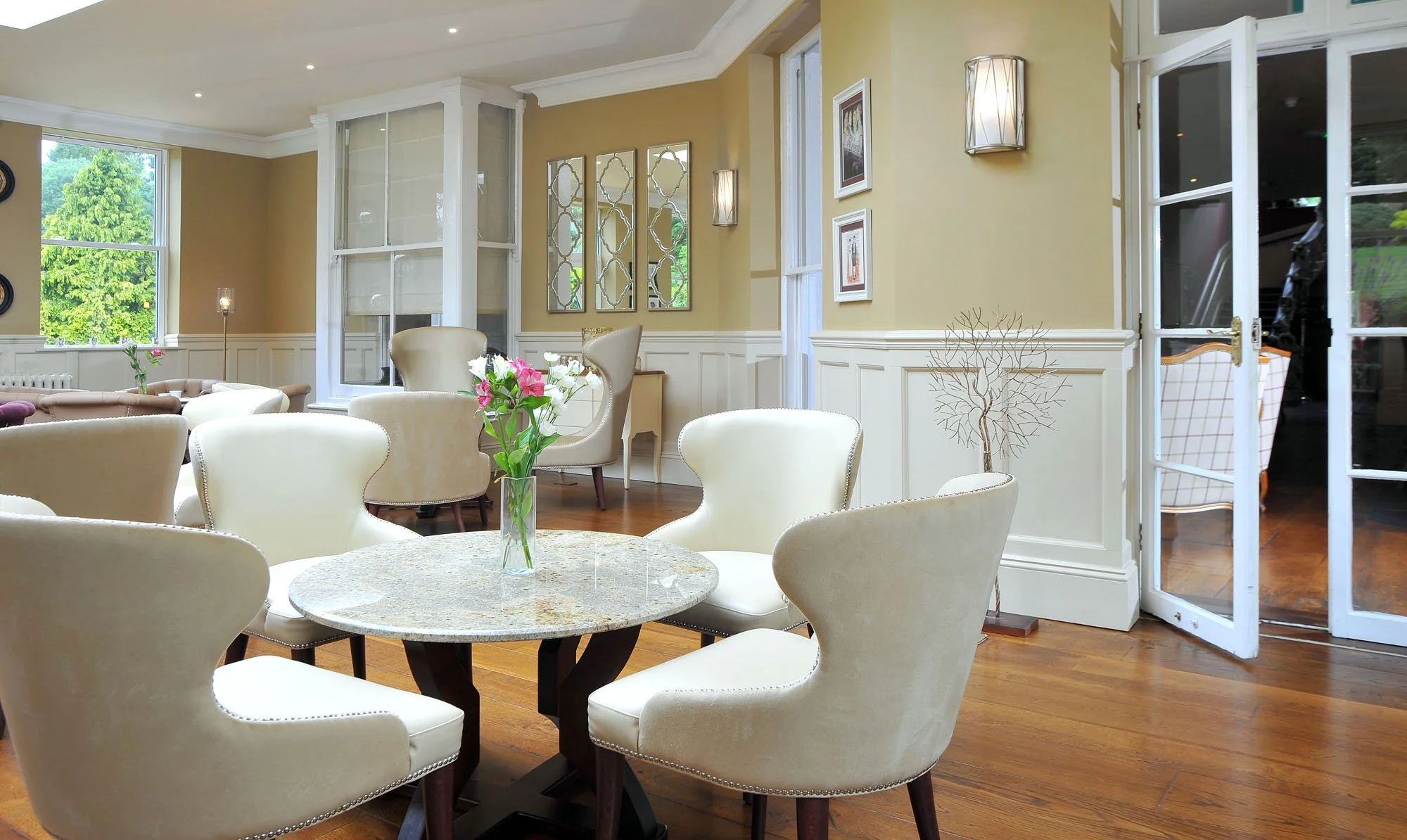 Bright dining room with a round table, cream-colored chairs, and a vase of pink and white flowers. Large windows show a green outdoor scene, and there are wall lights and framed artwork.