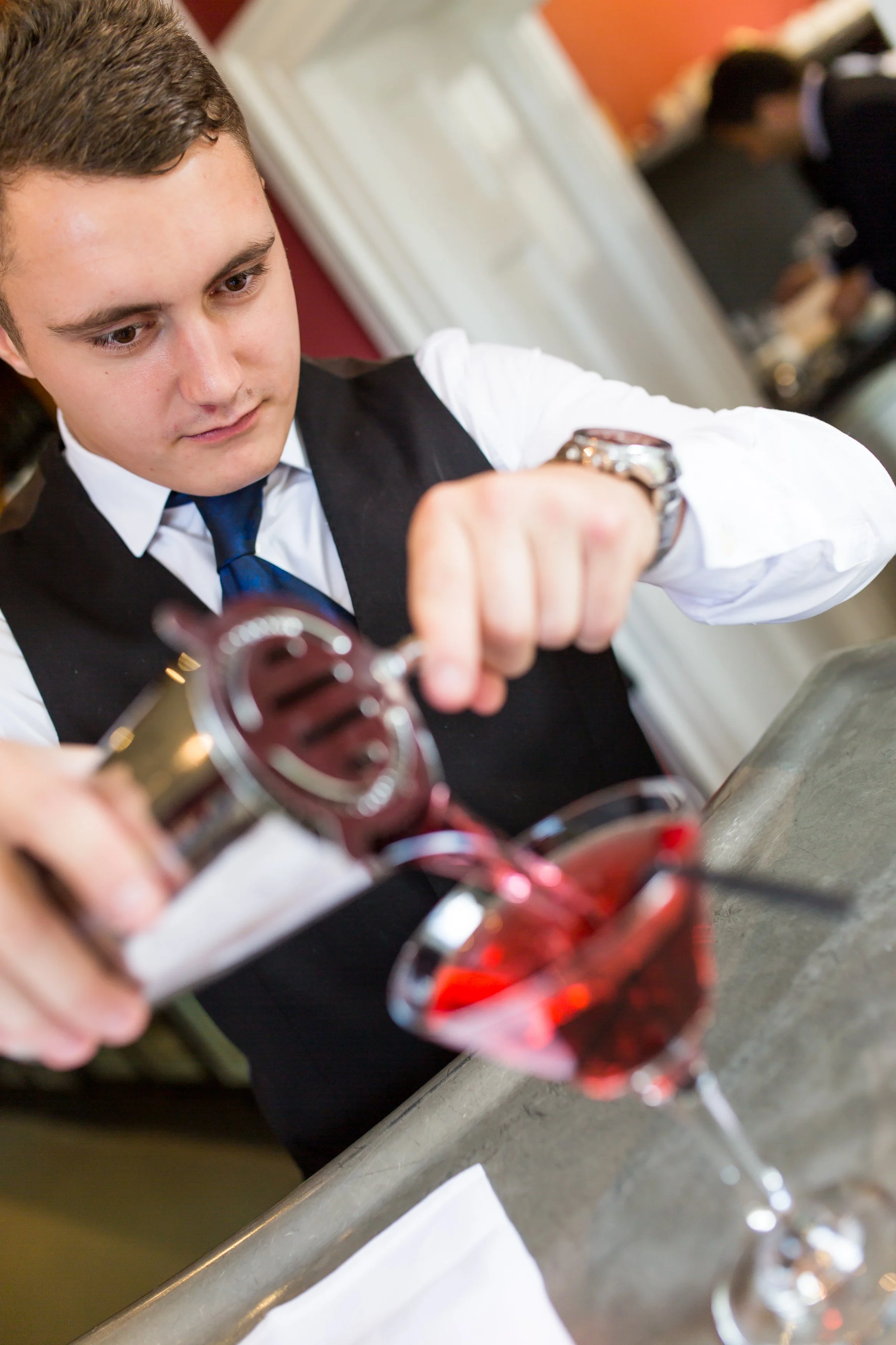 A young man in formal attire pouring red wine into a glass.