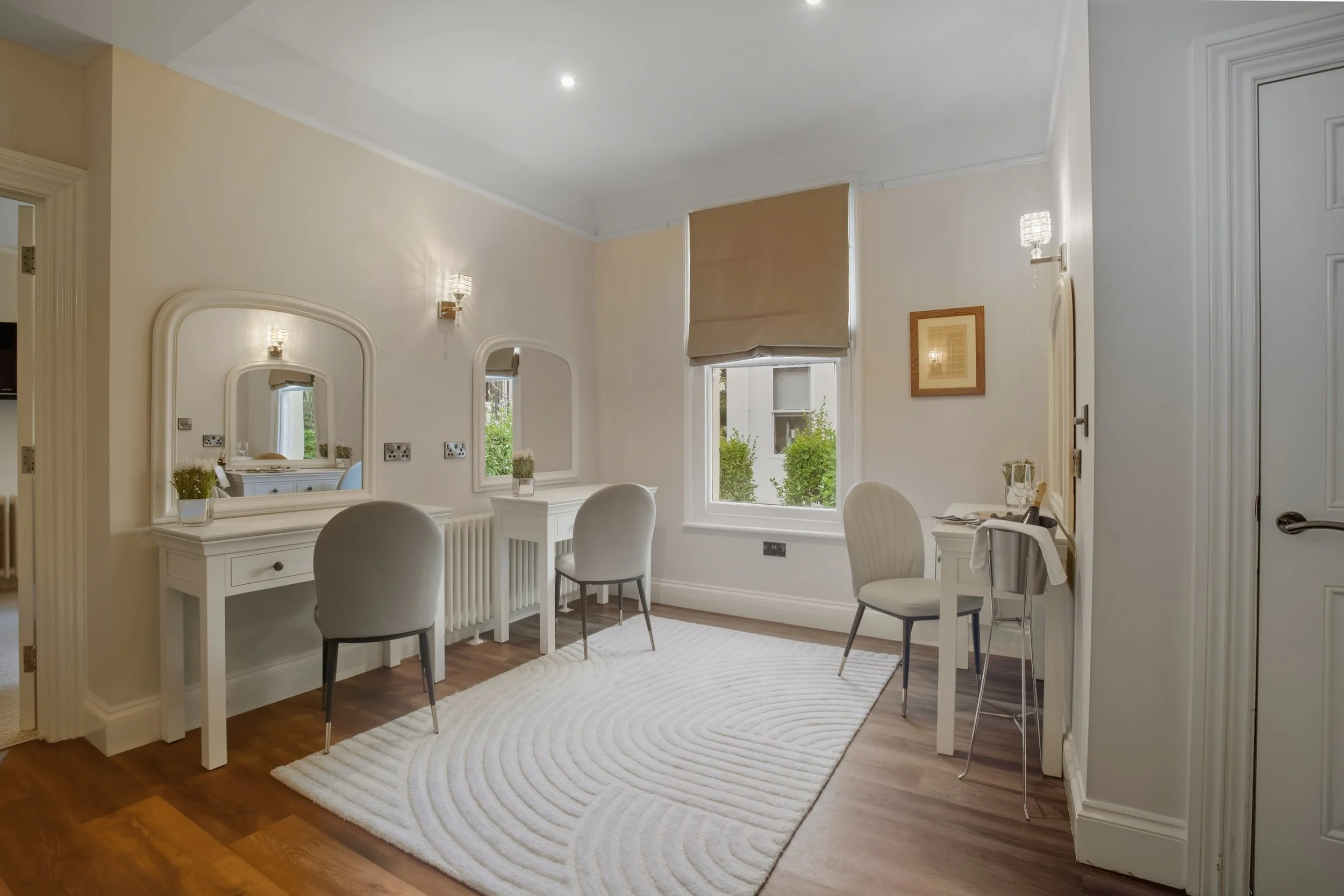 A bright, elegant room with a white vanity table, three chairs, a large window with a beige blind, framed artwork, and a white rug on wooden flooring.