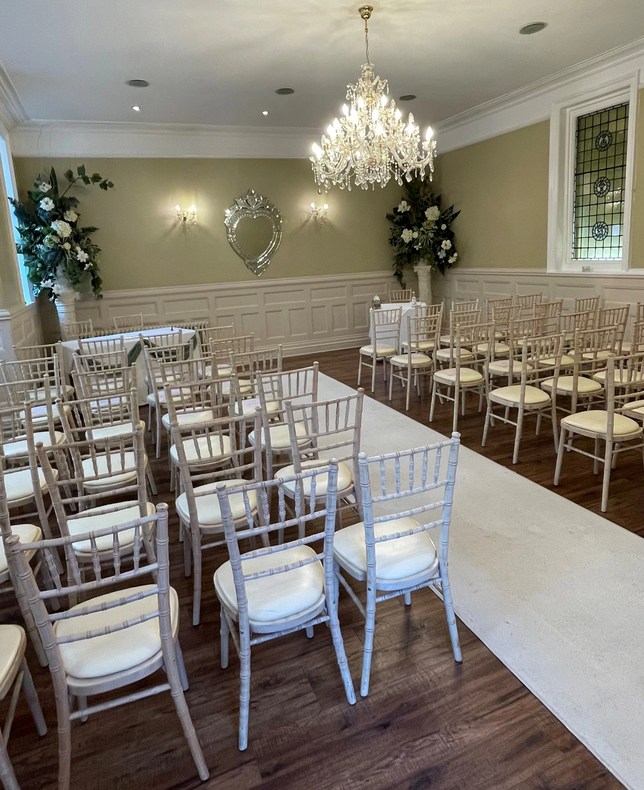 An elegant, decorated room set up for a wedding or special event, with white chairs, floral arrangements, a chandelier, and a white aisle runner.