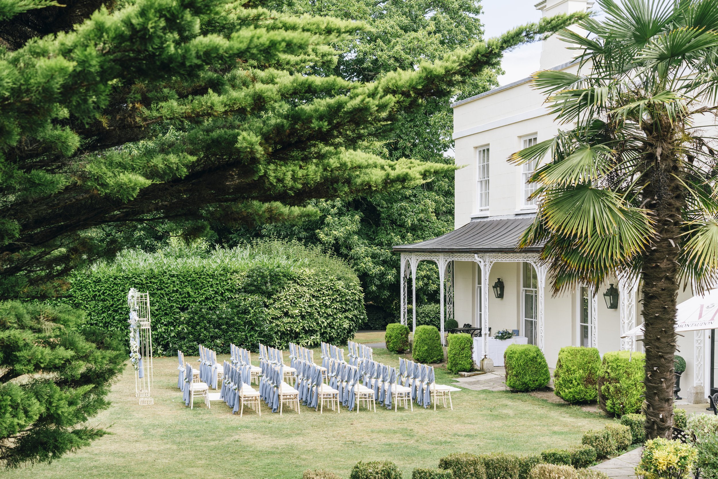 Outdoor wedding setup with rows of chairs draped in light blue fabric, positioned on green grass in front of a white house with decorative porch columns, surrounded by lush green trees and bushes.
