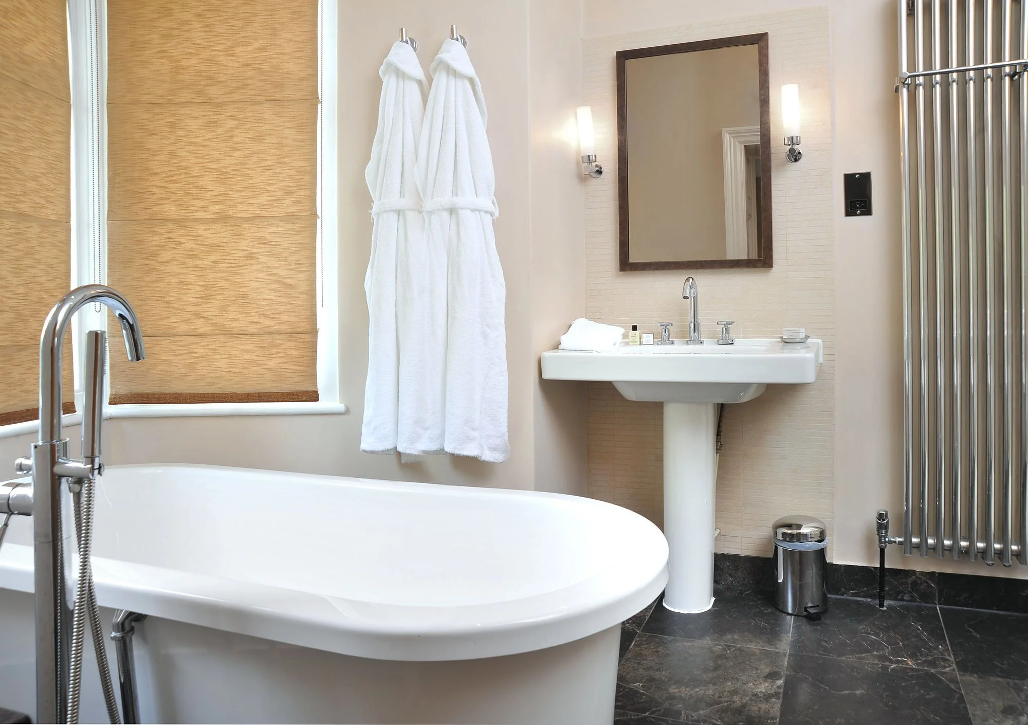 Bathroom with a bathtub, white towels hanging on wall, a mirror with two wall sconces, a sink with toiletries, a window with a bamboo shade, and a chrome radiator.
