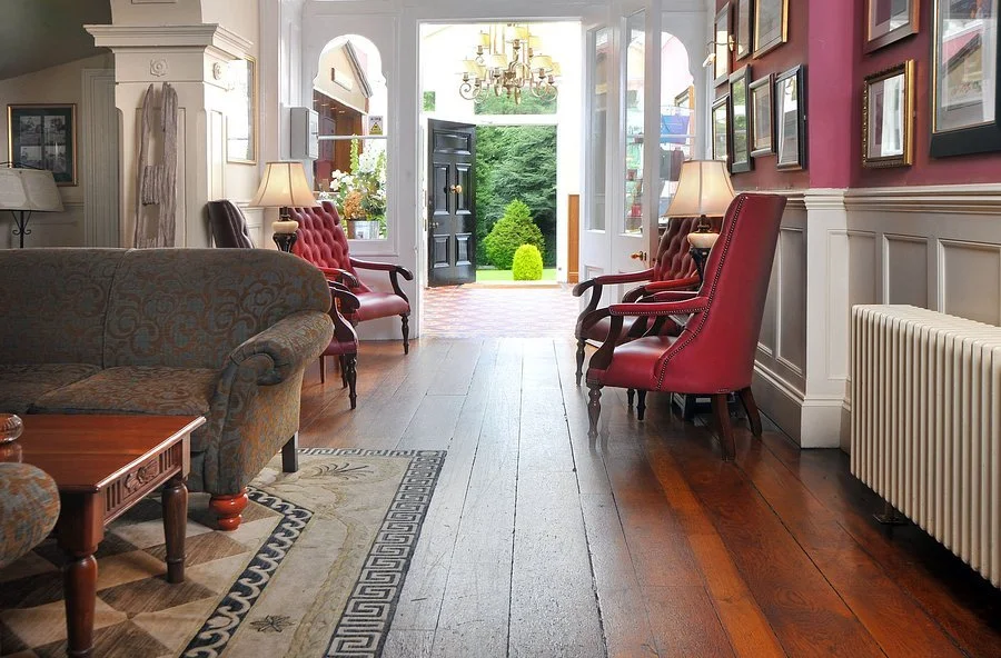 Entrance hall with wooden flooring, red armchairs, lamps, framed pictures on a red wall, and a view of a green garden outside through an open door.